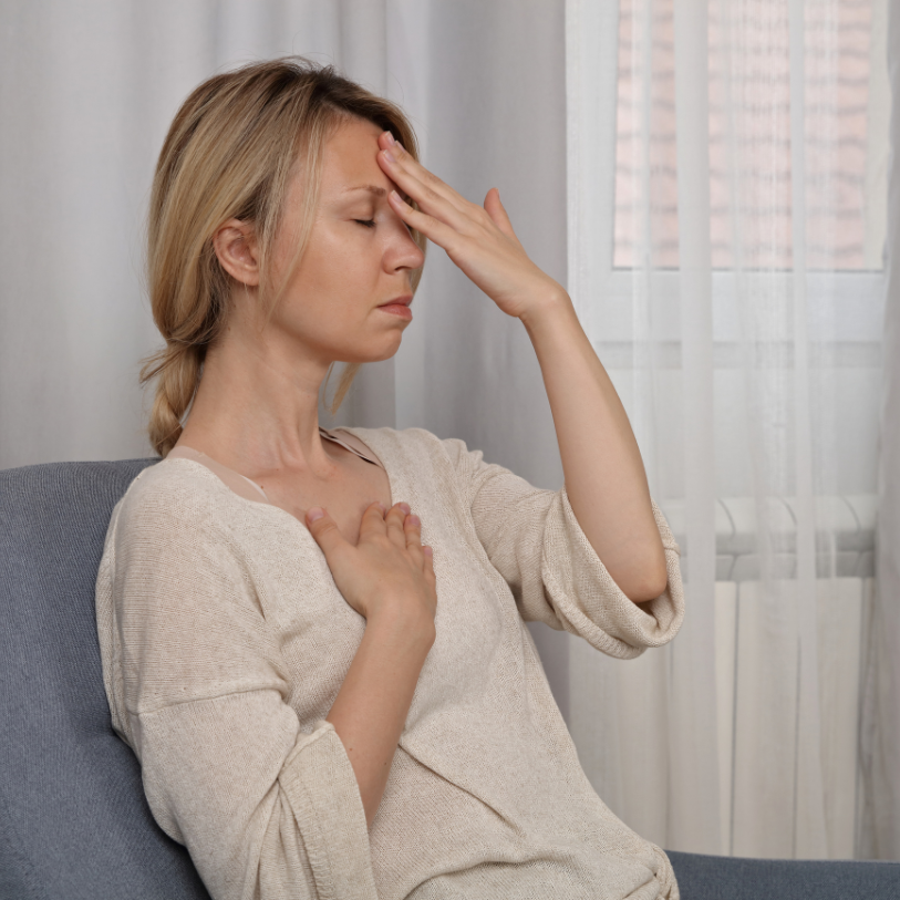 A woman sitting on a couch with her eyes closed, touching her forehead with one hand and her chest with the other, appearing to be experiencing discomfort or pain.