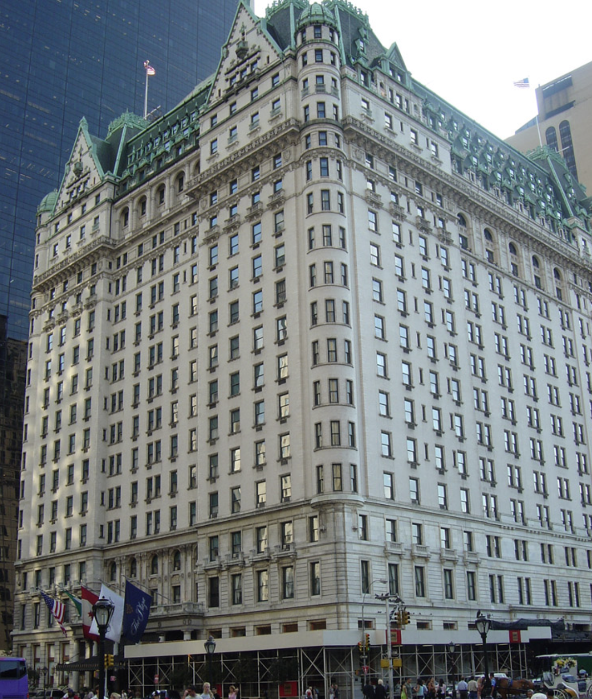 A tall historic building with a green mansard roof and white stone facade, situated in an urban area with people and flags at street level.