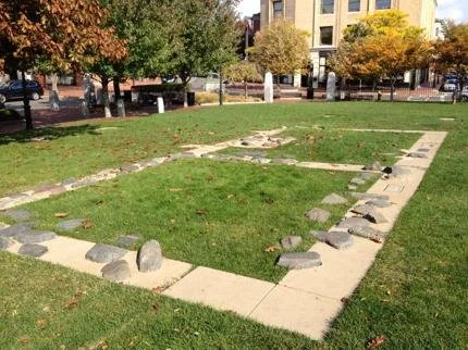 Remains of a rectangular structure with concrete blocks and scattered rocks in a park with trees and buildings in the background.