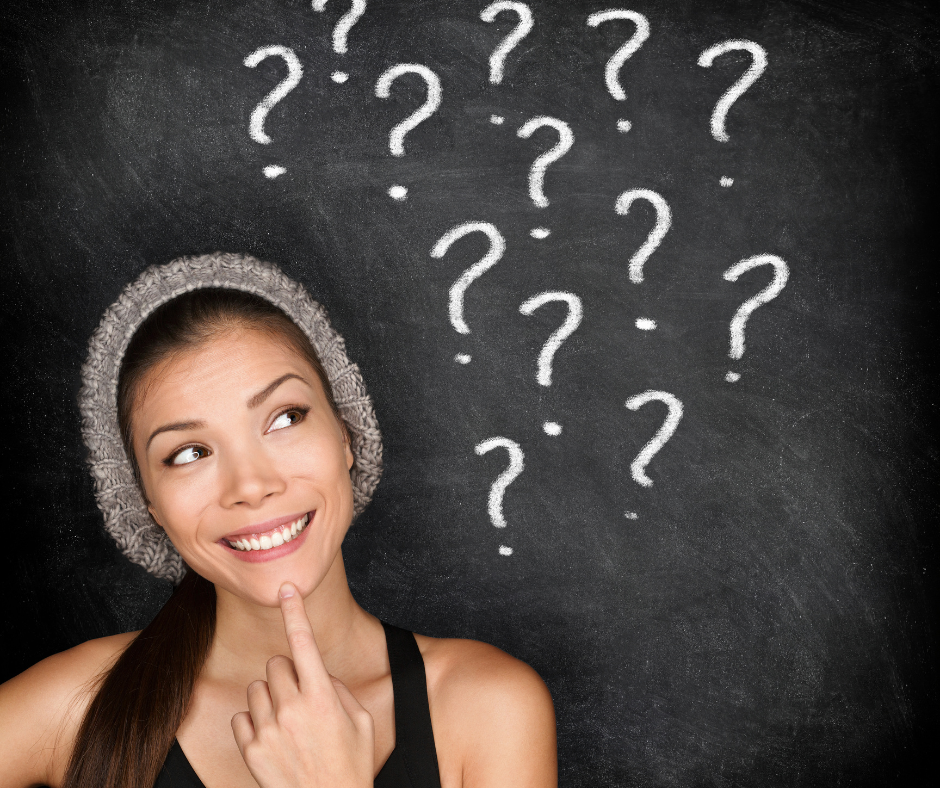 A woman with a thoughtful expression, touching her chin, standing in front of a blackboard with multiple chalk-drawn question marks.