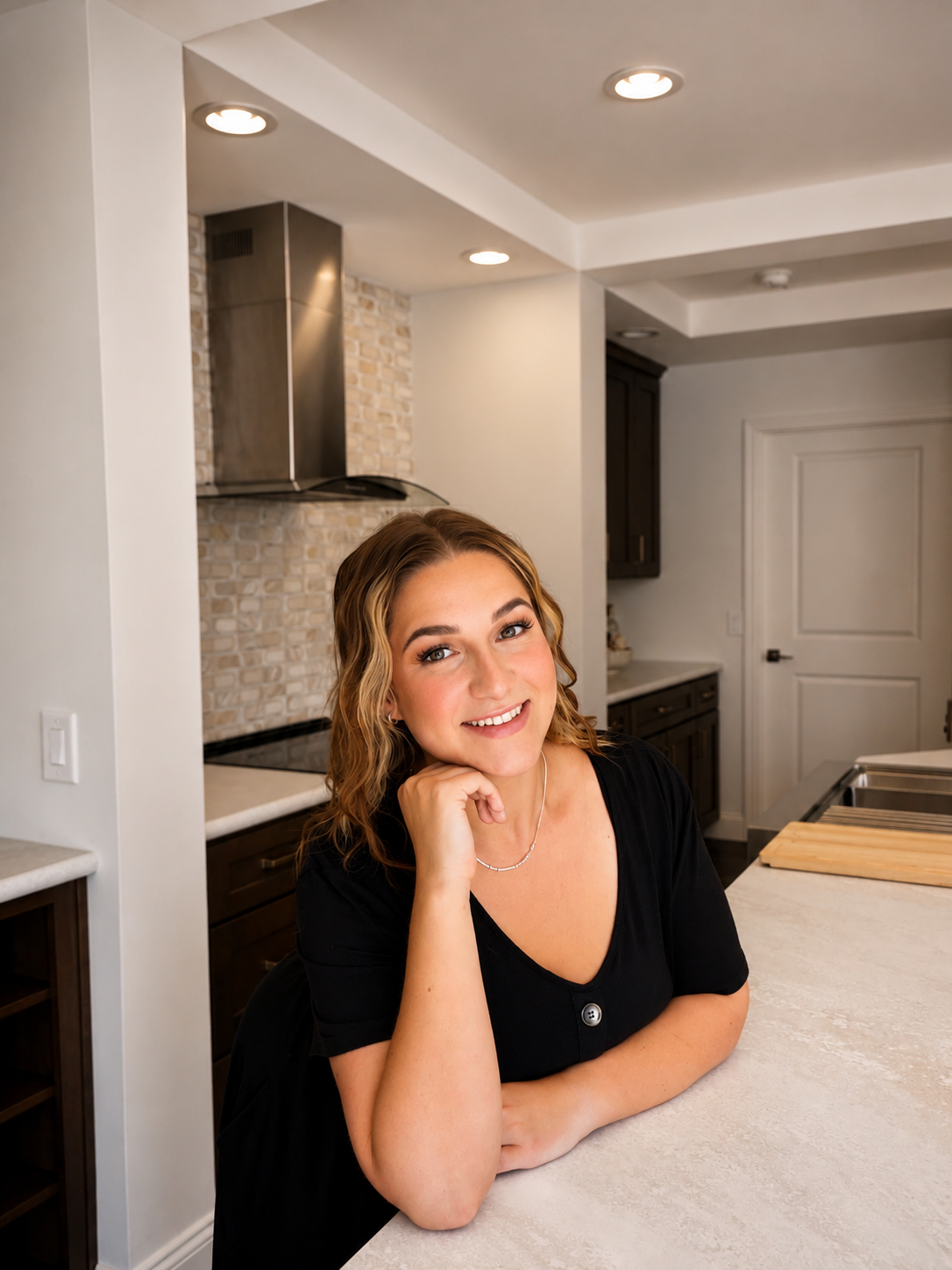 A smiling woman with curly hair, wearing a black top, sitting at a kitchen counter with a beige countertop. The kitchen has modern design elements with dark cabinets, a brick backsplash, and stainless steel appliances.