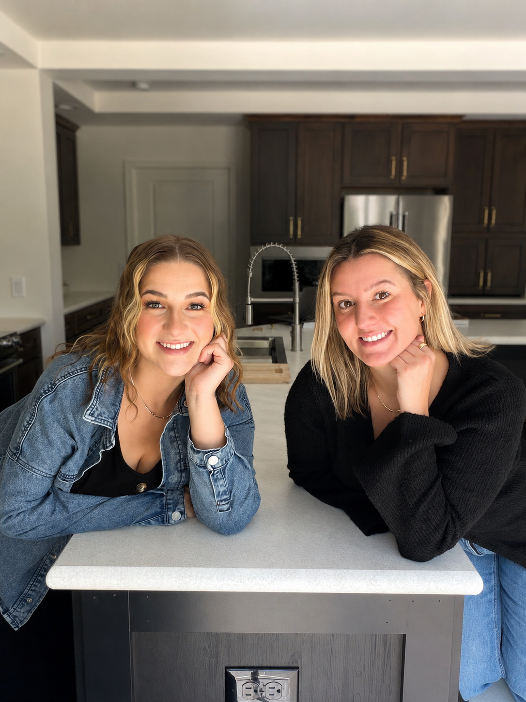Two young women with wavy blonde hair smiling and leaning on a white kitchen island, with dark cabinets and stainless steel appliances in the background.