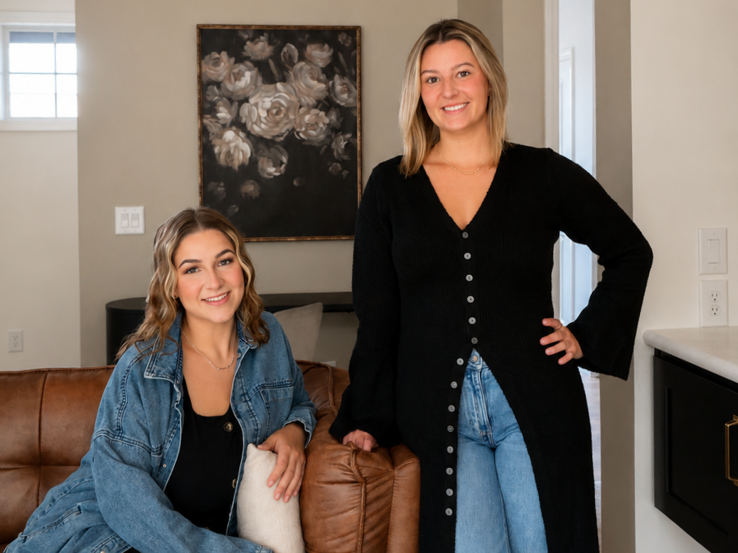 Two young women smiling in a living room with neutral-colored walls, a leather sofa, and an abstract floral painting behind them. One is sitting on the sofa with a denim jacket, and the other is standing with her hand on her hip, wearing a black cardigan and blue jeans.