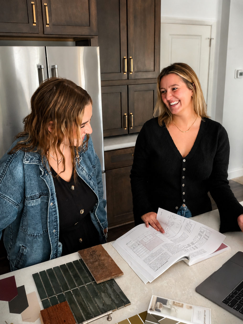 Two women standing at a kitchen counter looking at tile and design samples and a large open design book, smiling and engaged in conversation.