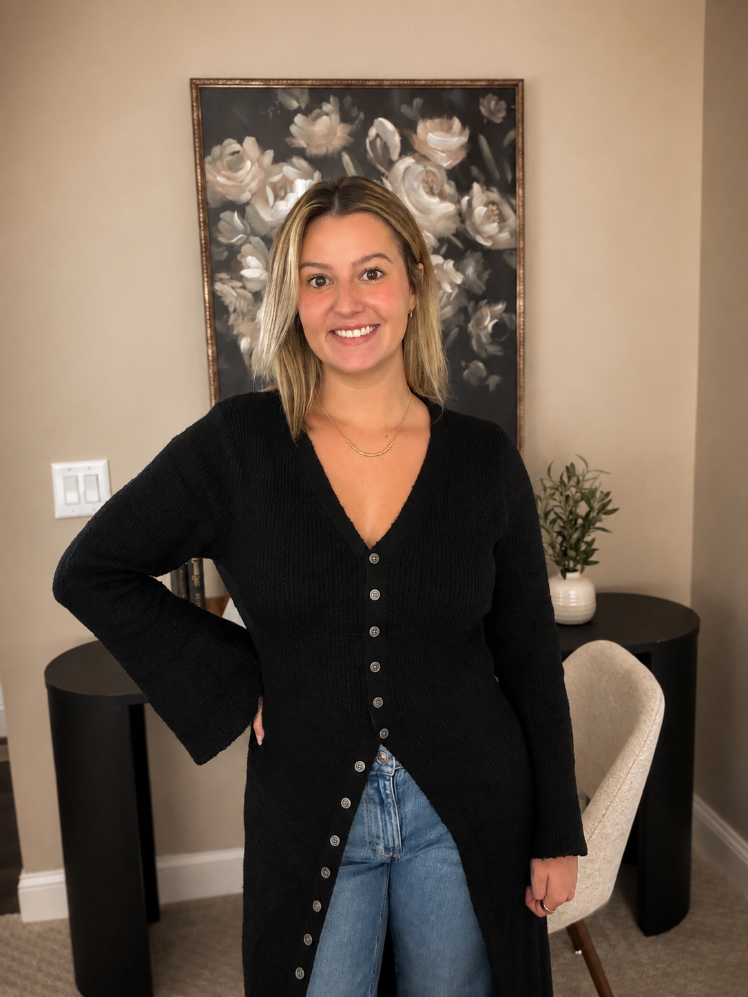 A young woman with shoulder-length blonde hair, wearing a black long cardigan and blue jeans, smiling and standing indoors near a beige wall with a large flower painting and a small table with a potted plant.