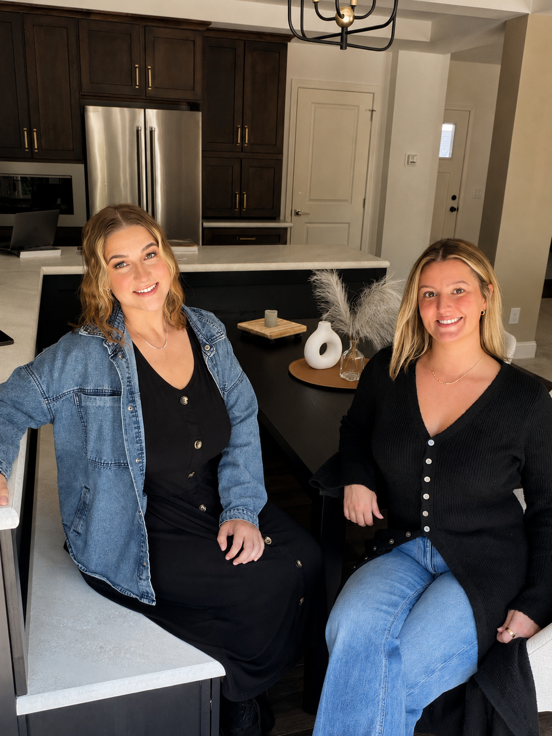 Two women sitting in a modern kitchen, smiling at the camera. The kitchen has dark wood cabinets, a stainless steel refrigerator, and minimalist decor with white vases and pampas grass on the table.