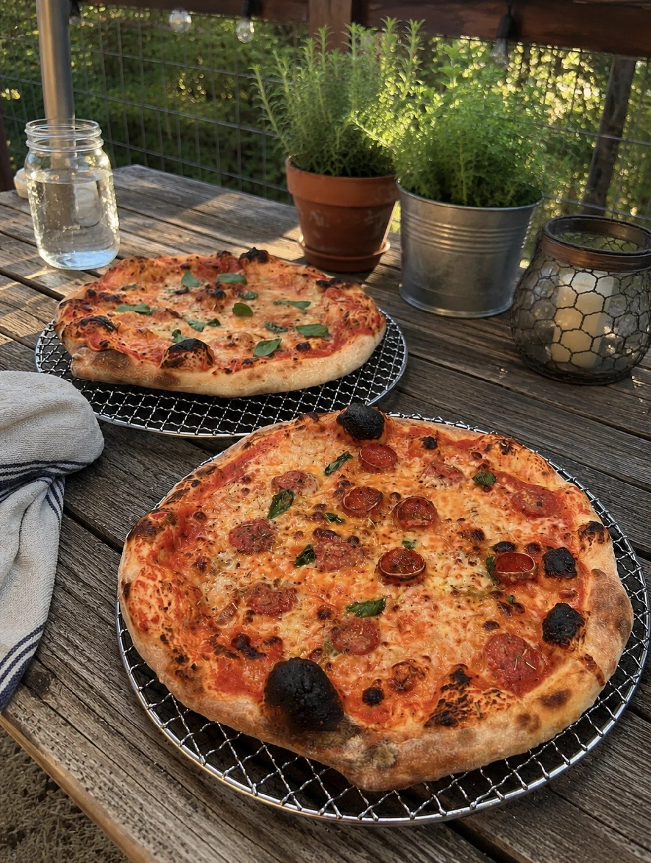 Two pizzas on a picnic table outdoors, with potted plants and candles in lanterns nearby, and a jar of water