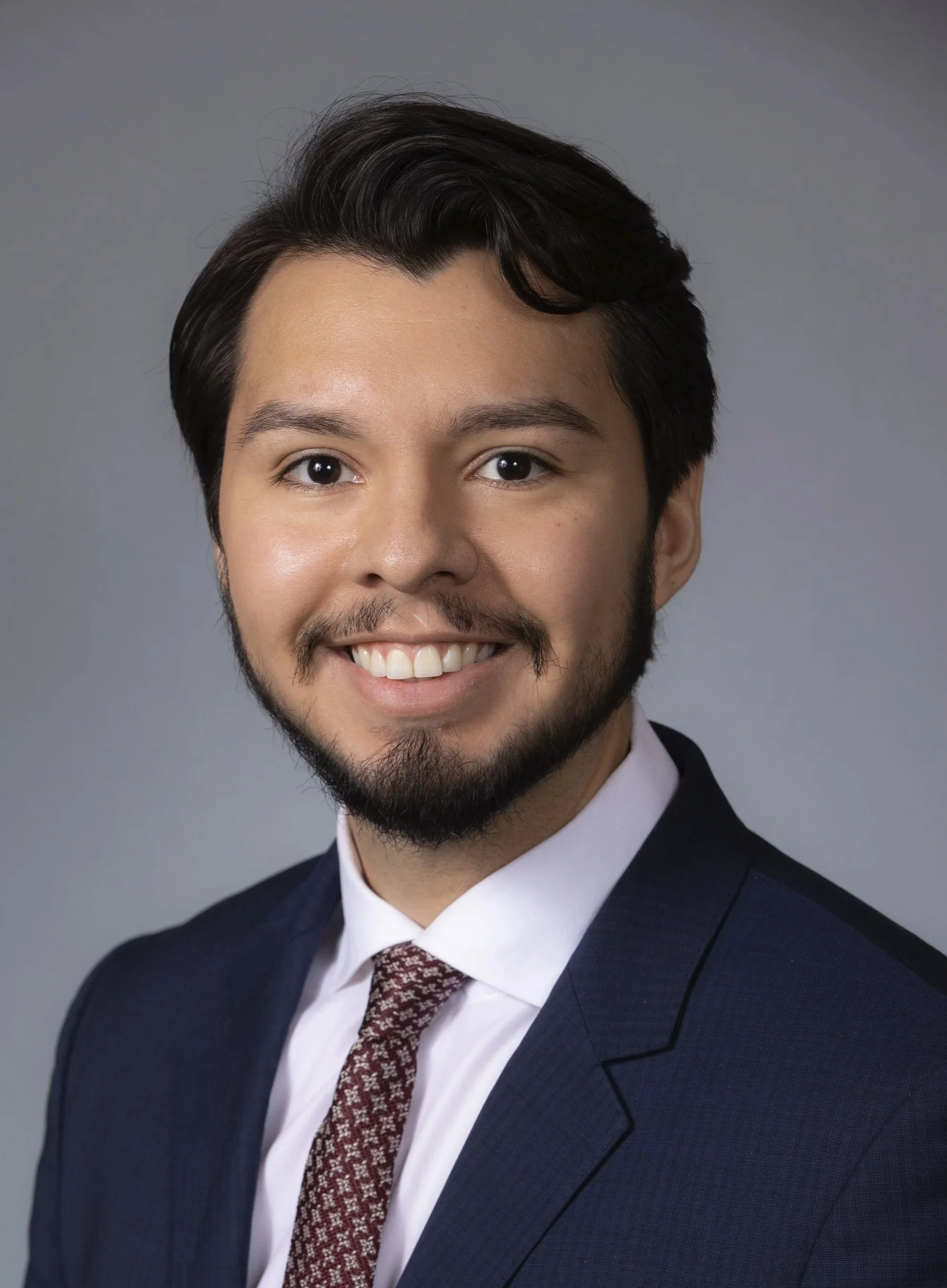 Close-up of a smiling young man with dark hair, beard, wearing a navy suit, white shirt, and patterned tie, against a gray background.