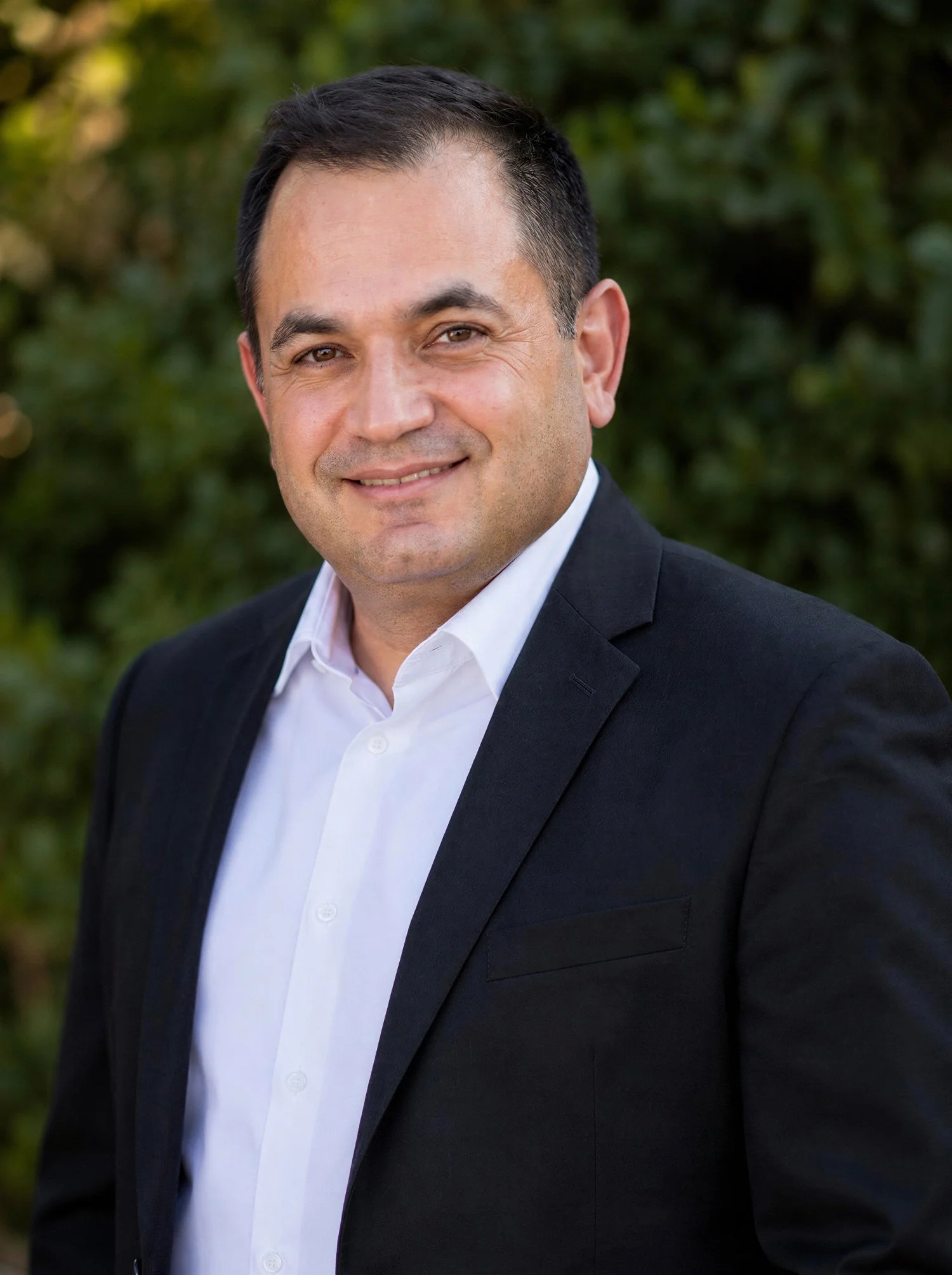 A man in a black suit and white shirt smiling outdoors with a blurred green tree background.