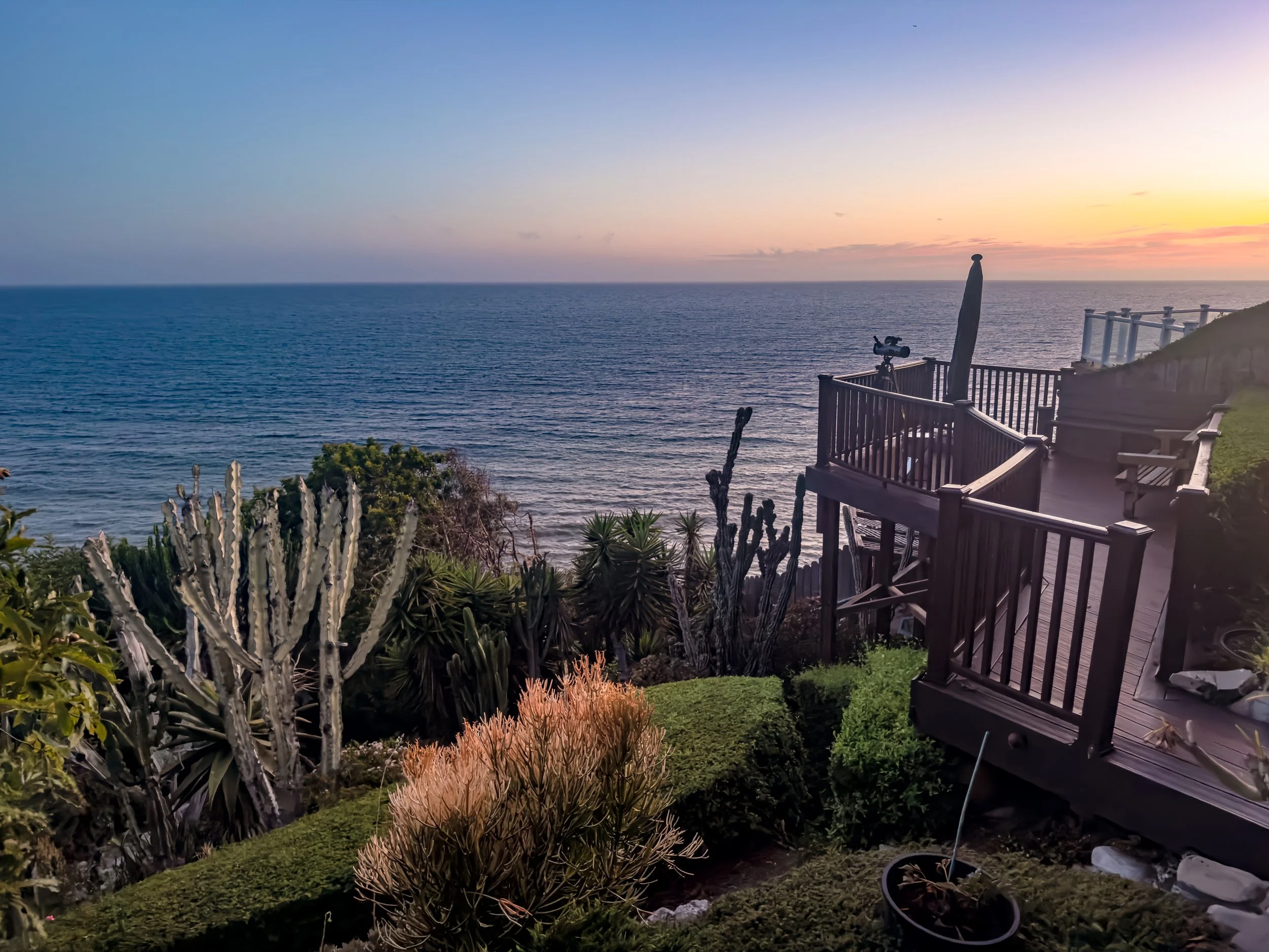 A scenic view of the ocean at sunset with a wooden deck, telescope, and various plants in the foreground.