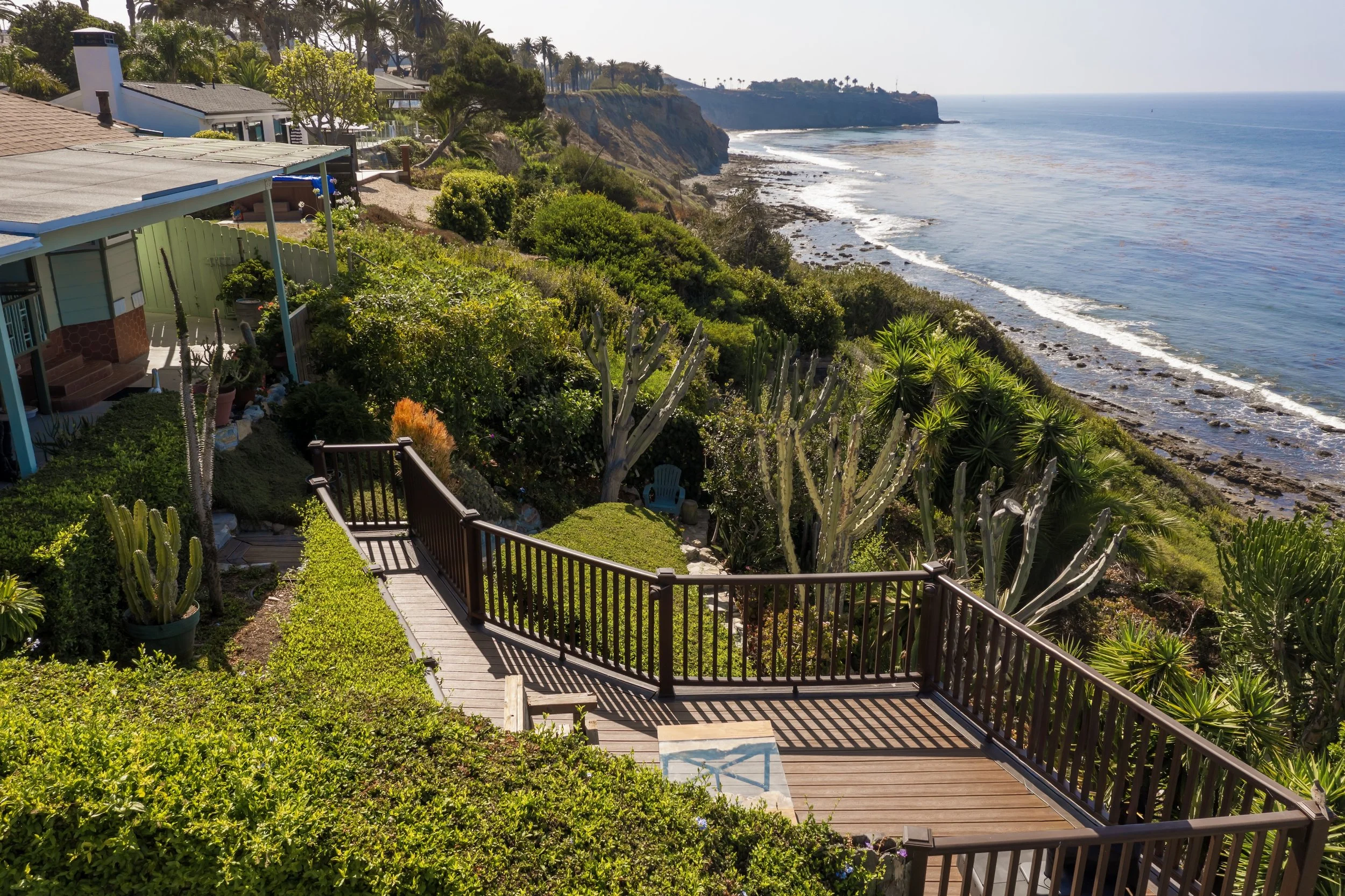 A view of ocean cliffs with houses and lush greenery, including cacti, along a shoreline with rocky and sandy beach. Wood and rail fencing on a garden patio.
