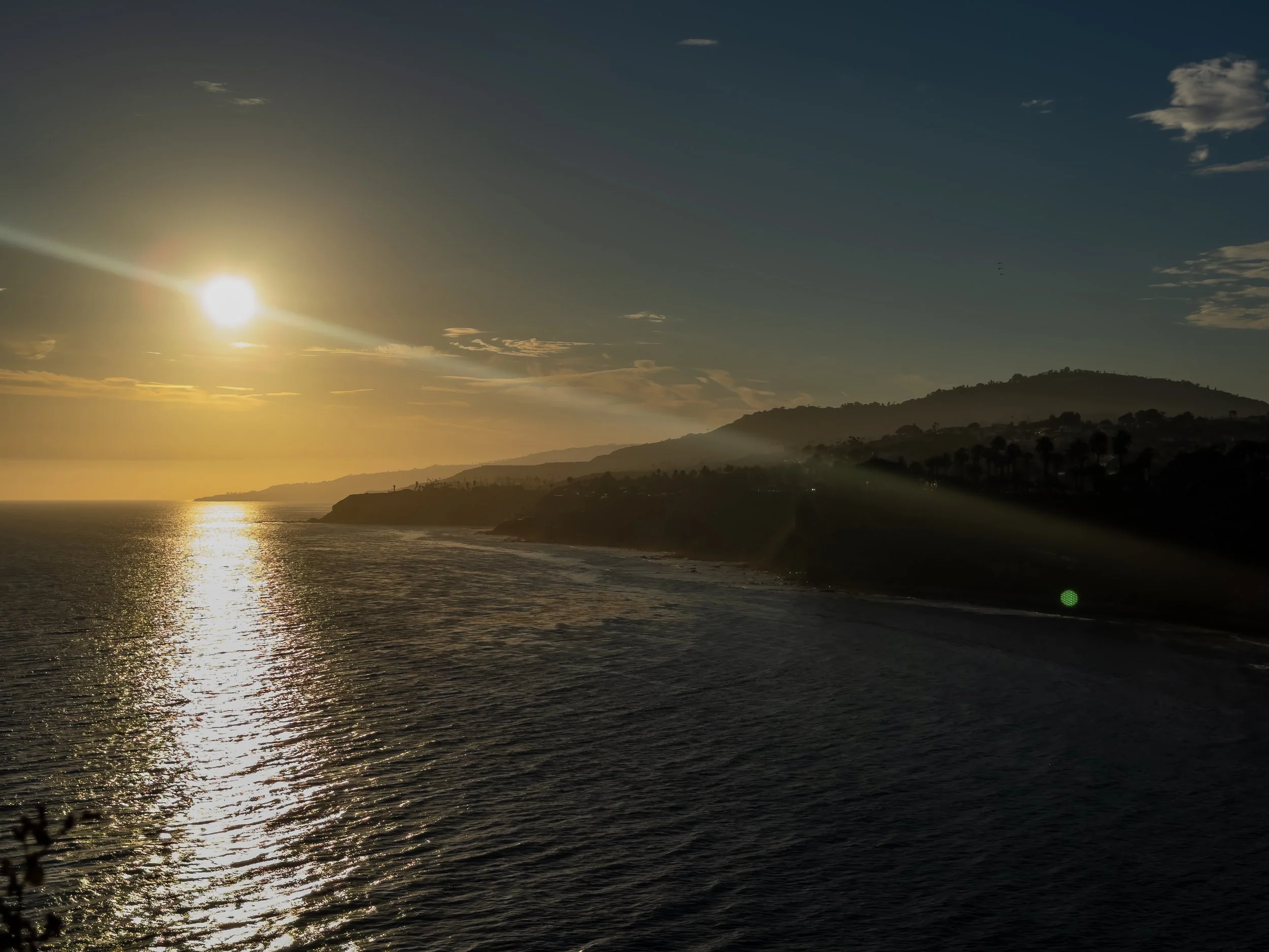 A scenic view of the ocean with a coastline, hills, and trees in the distance, under a sunset sky with some clouds.