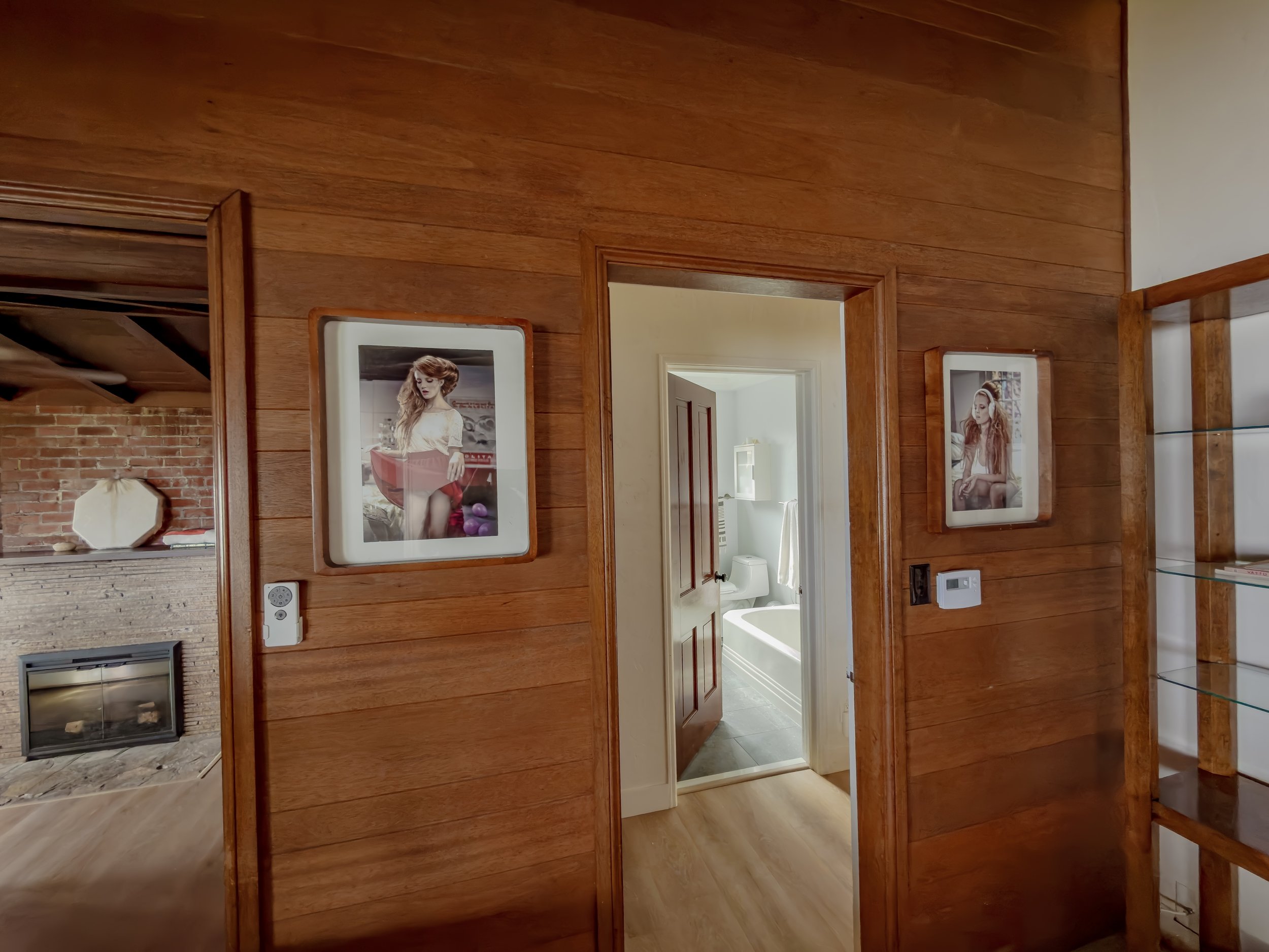 Interior view of a wooden wall with framed photographs, a doorway leading to a bathroom, and a fireplace in the background.
