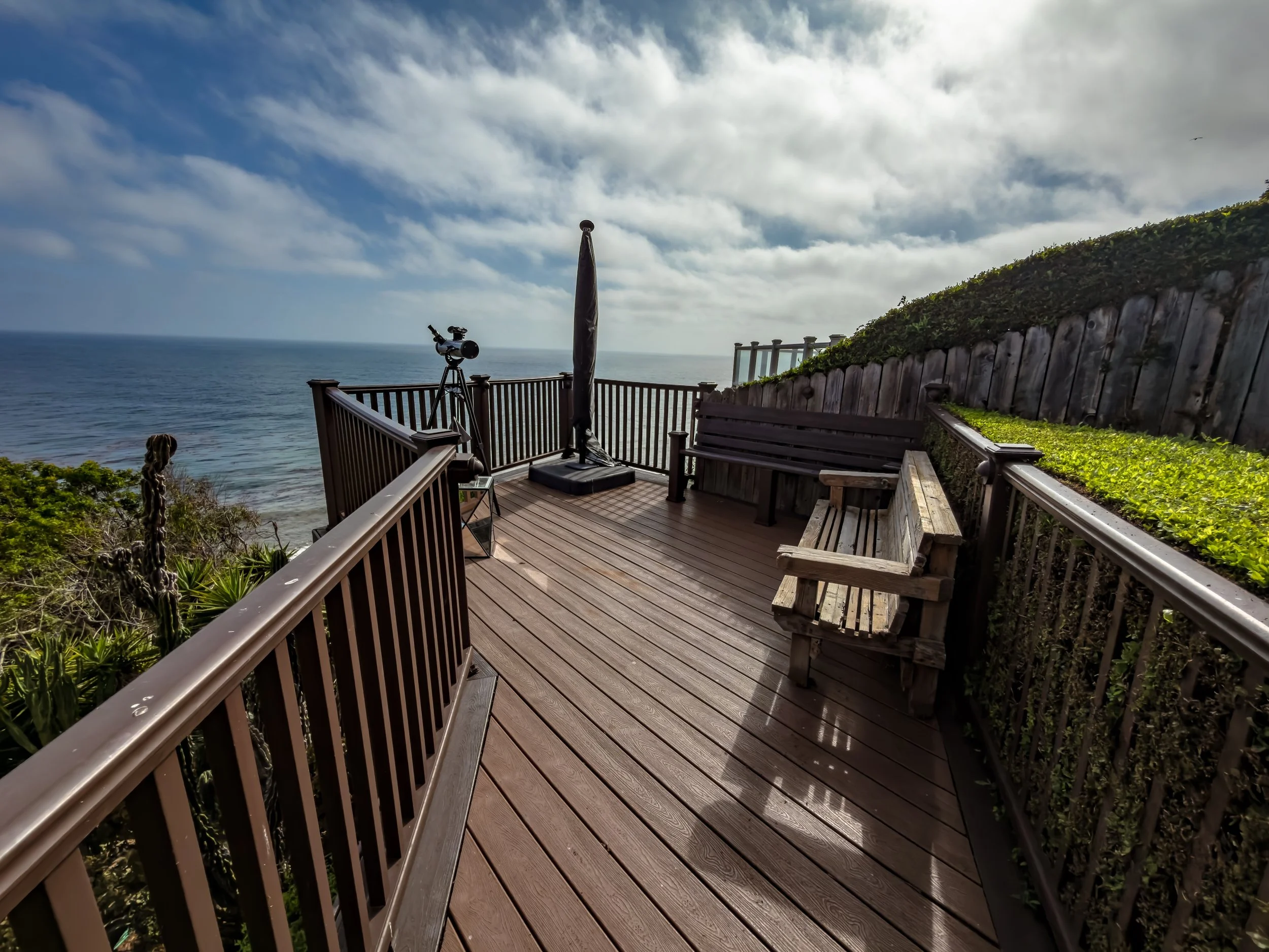 A wooden deck overlooking the ocean with a telescope, benches, and a black umbrella, surrounded by greenery and a fence, under a cloudy sky.