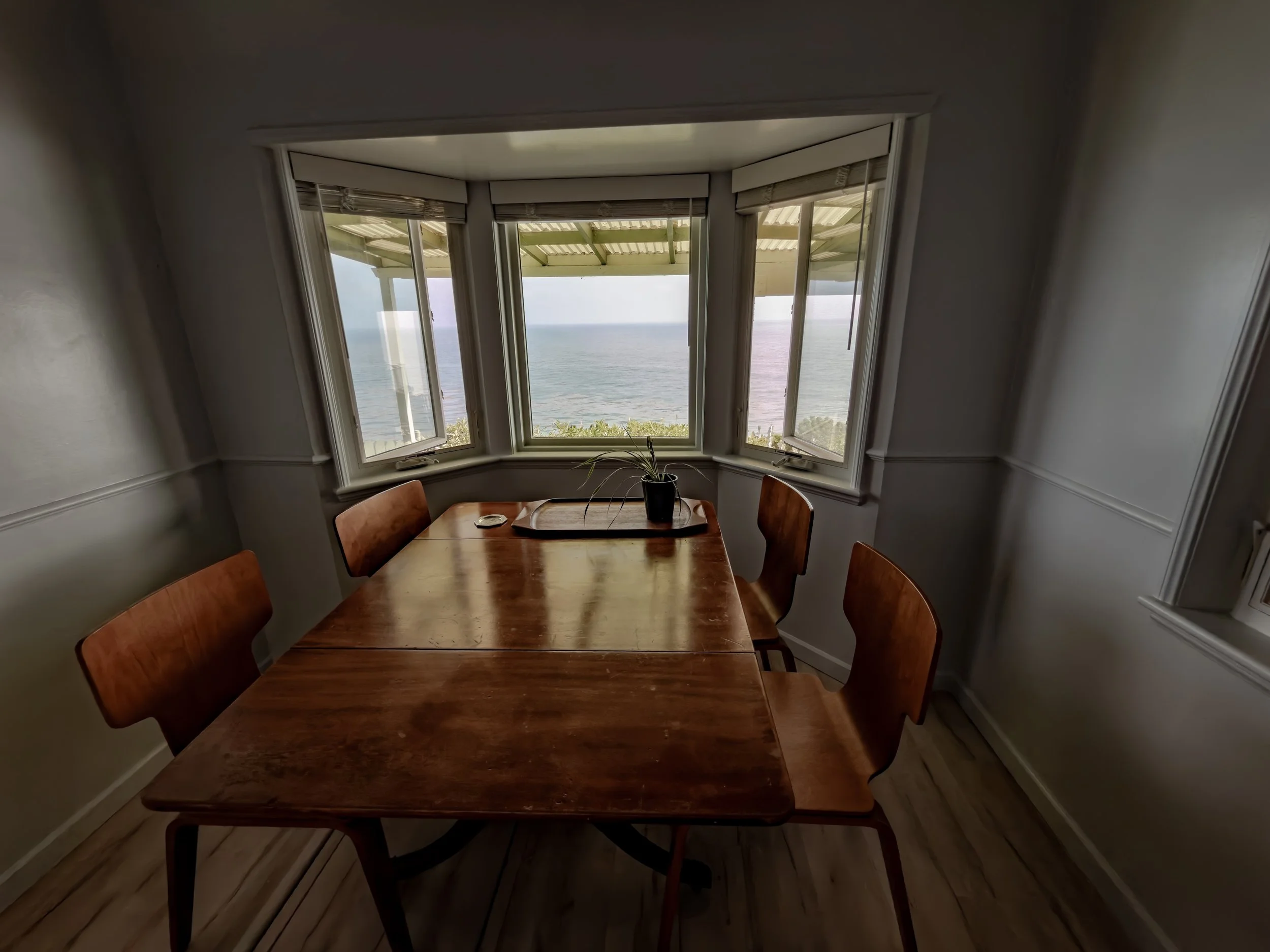 A wooden dining table with four chairs around it, placed near large windows overlooking the ocean and sky, with a potted plant on the table.