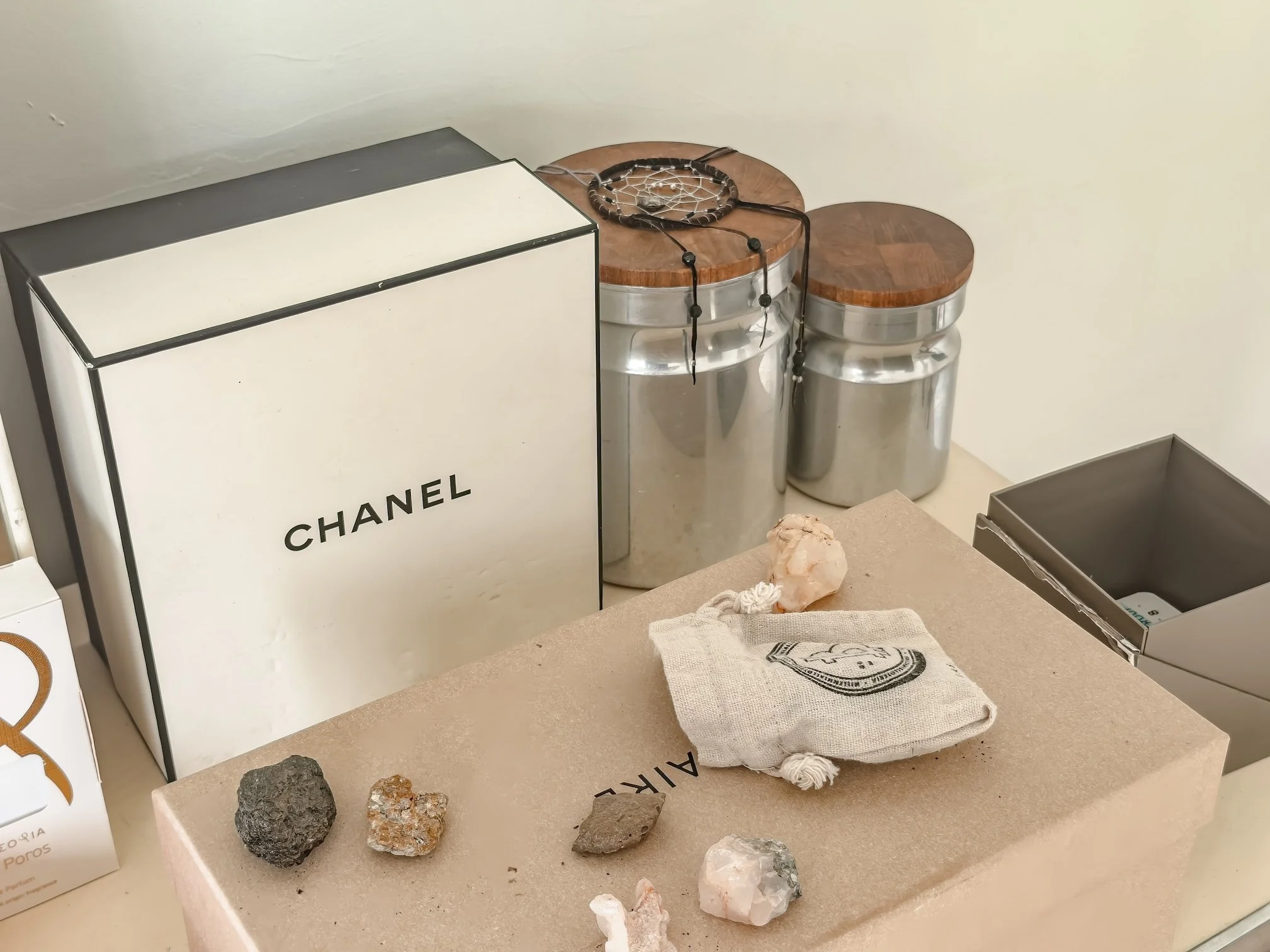 An educational display showing various rocks and minerals on a beige table. A white box with the word 'CHANEL' partially visible is on the left. There are two stainless steel containers with wooden lids towards the back. In front, there are several d