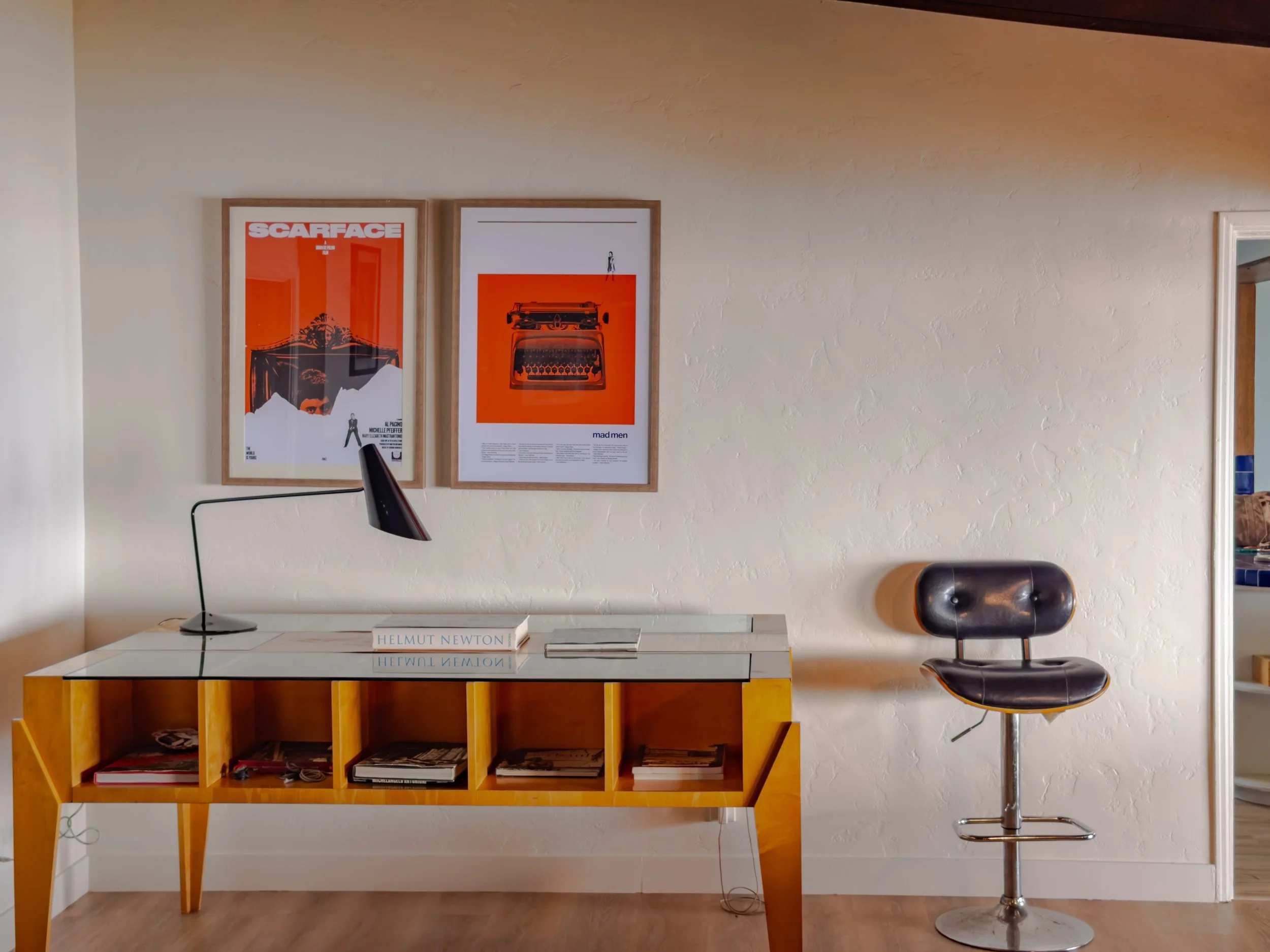 A modern room with a white textured wall and a wooden desk with a glass top. On the wall are two framed posters with orange backgrounds; one reads 'SCARFACE' and the other features a vintage camera illustration. Below the posters, there's a white boo