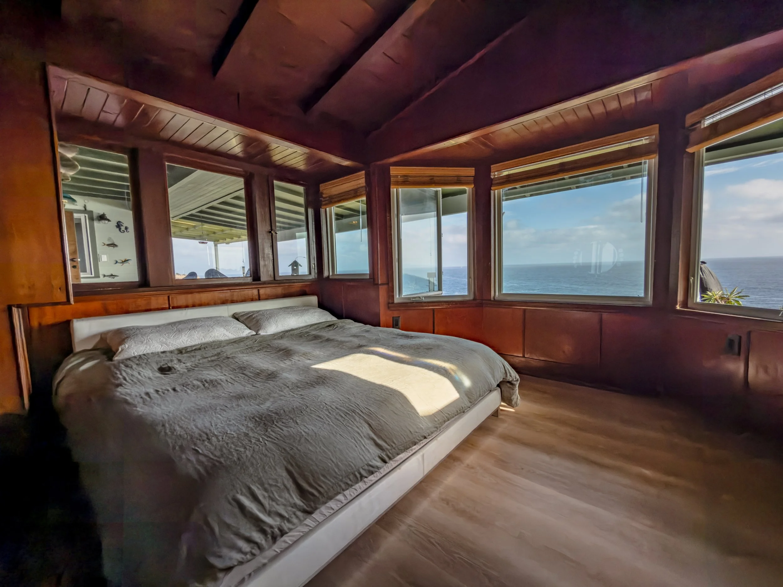 View of a bedroom with large windows overlooking the ocean, a bed with white bedding, wooden walls and ceiling, and partially opened blinds.