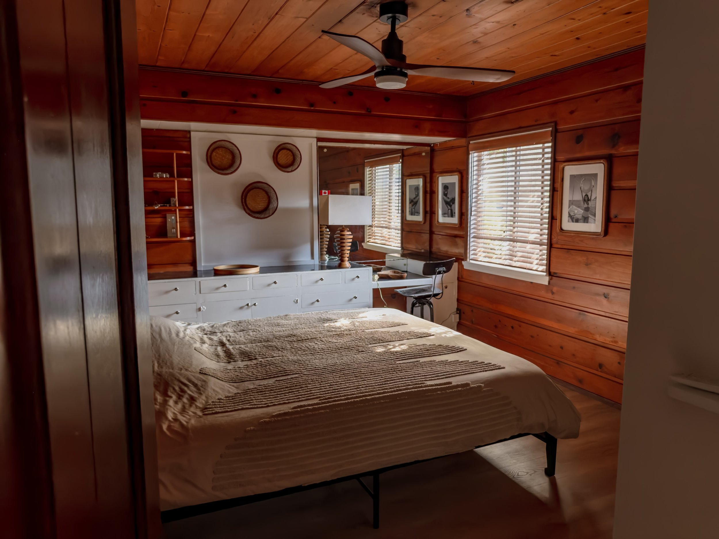 A cozy bedroom with wood-paneled walls and ceiling, featuring a bed with a beige bedspread, a dresser, and three framed black and white photographs on the wall. There are windows with blinds, a ceiling fan, and a hanging lamp near the window.