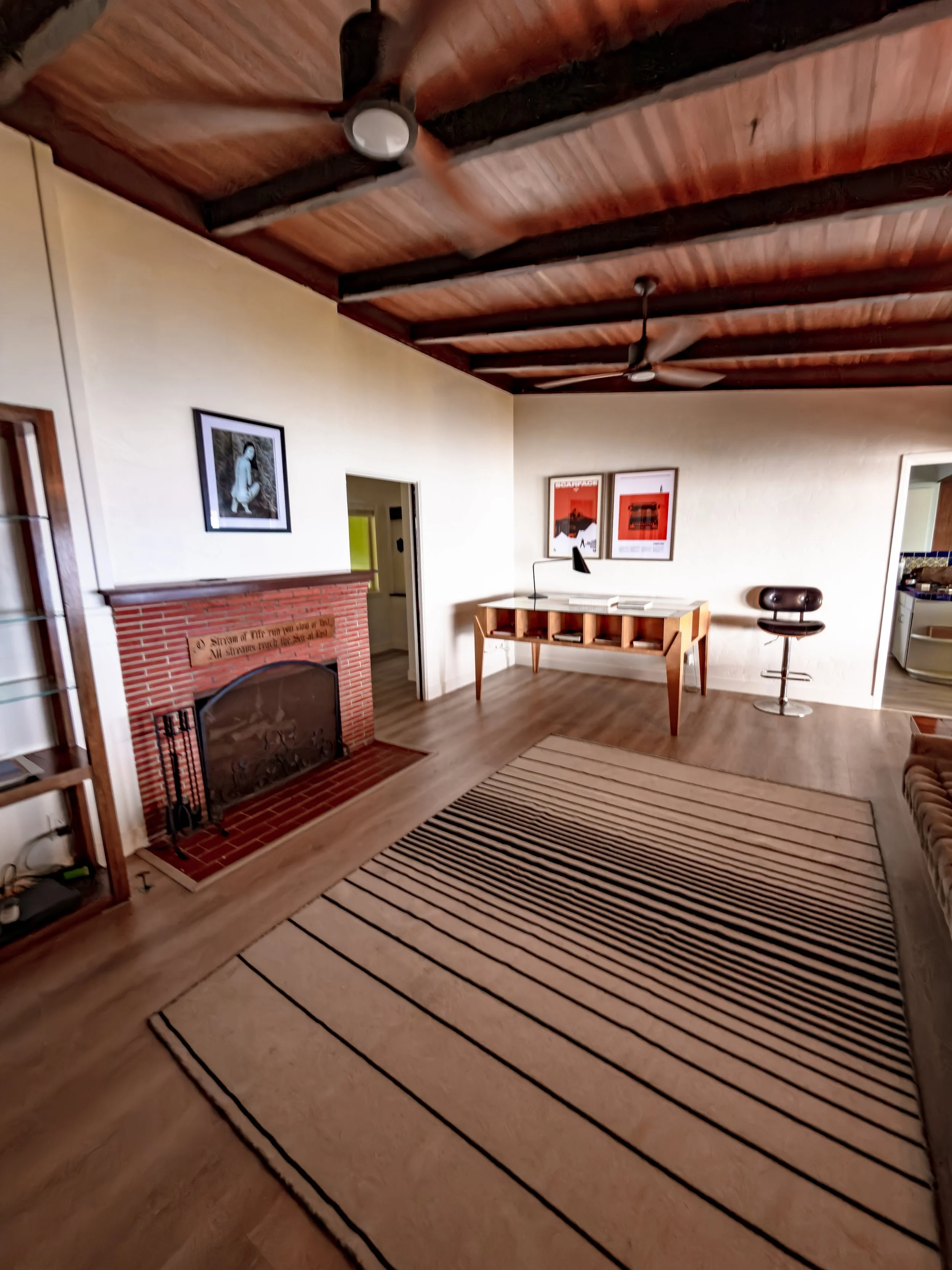 Living room with wooden ceiling, ceiling fans, artwork on the walls, fireplace, and a striped rug on wooden flooring.