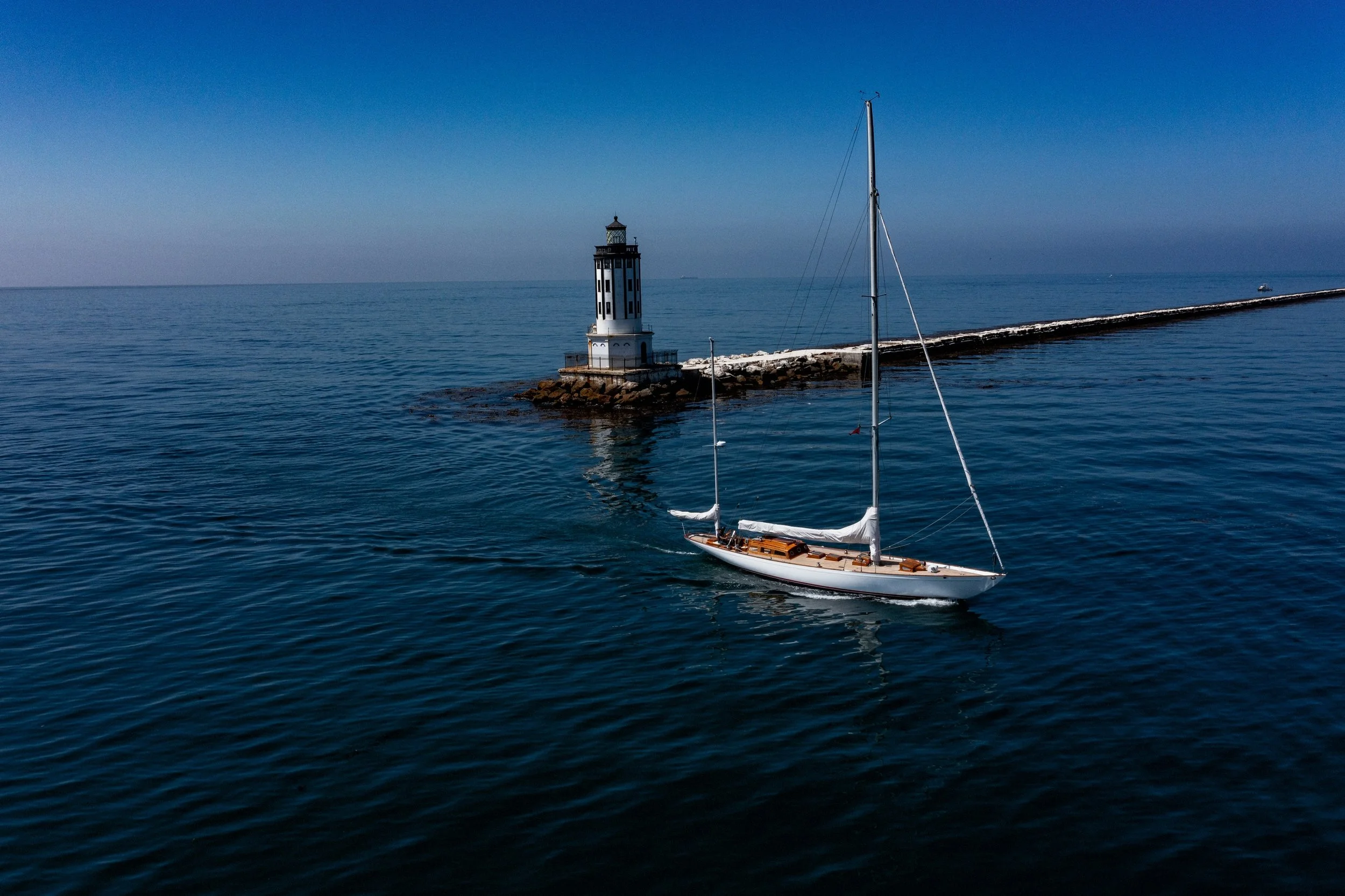 A sailboat in the water near a lighthouse on a rocky breakwater with a clear blue sky in the background.