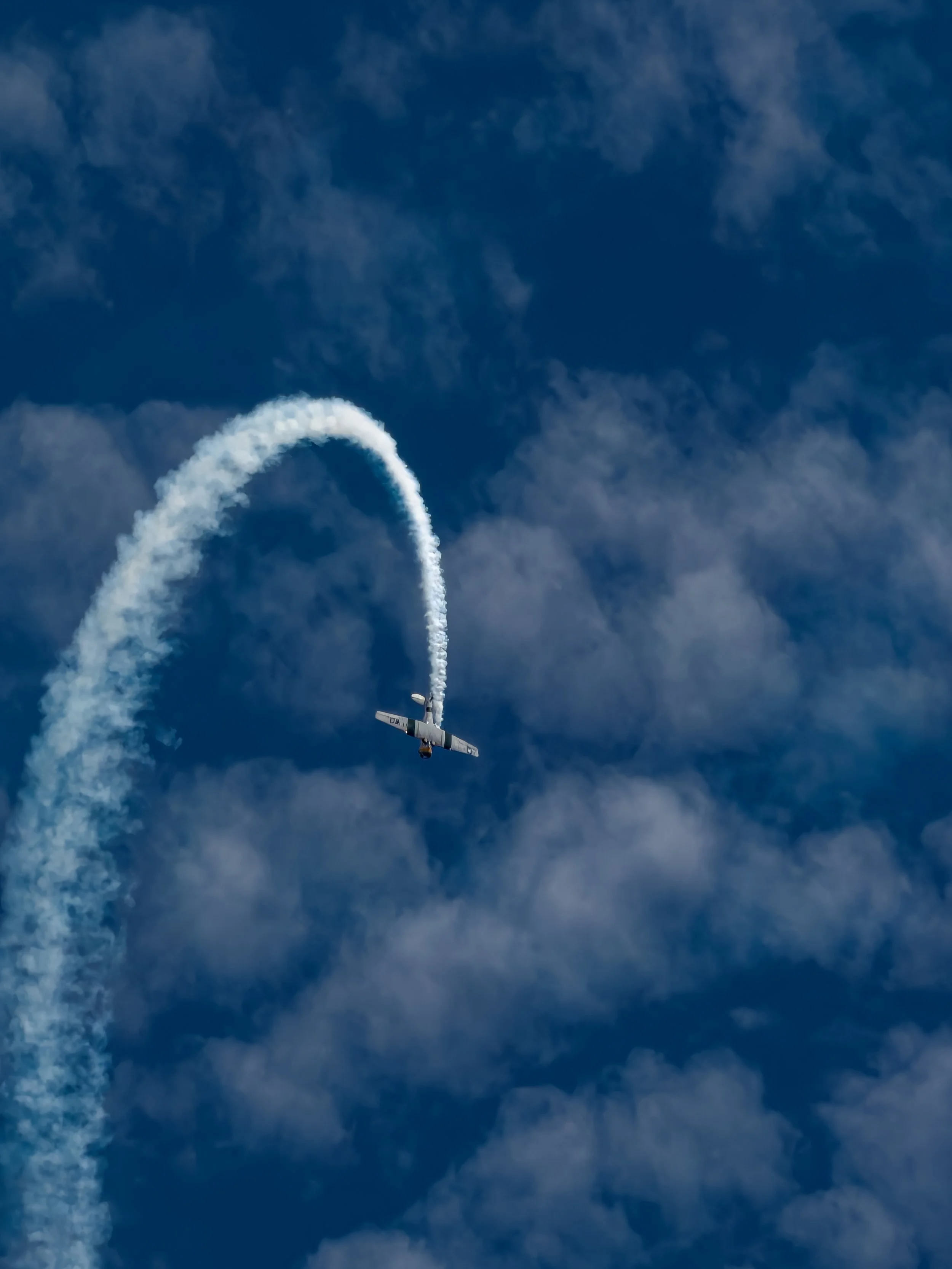 Small aircraft flying through the sky smoking a vapor trail in a loop shape against a partly cloudy blue sky.