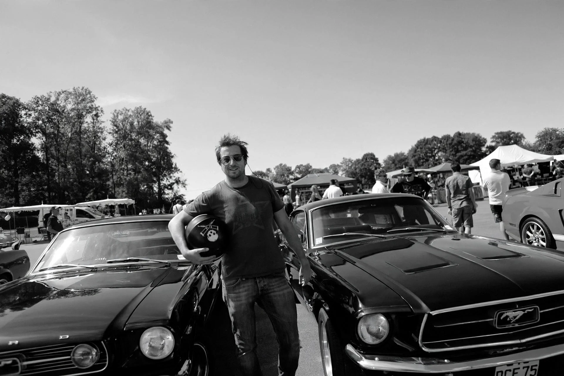 Jonathan Lennard  in sunglasses, holding motorcycle helmet, standing next to a vintage sports car at an outdoor car show, with tents and other people in the background.