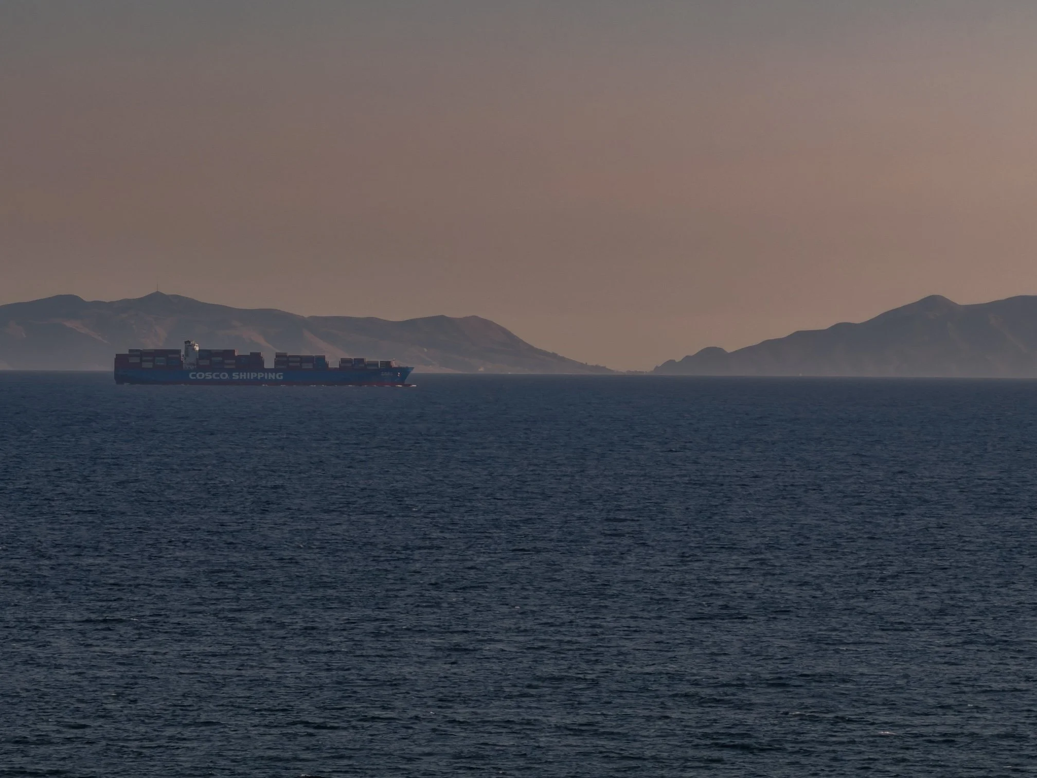 A large cargo ship labeled "COSCO SHIPPING" sailing on open water, with Catalina in the background under a clear sky.