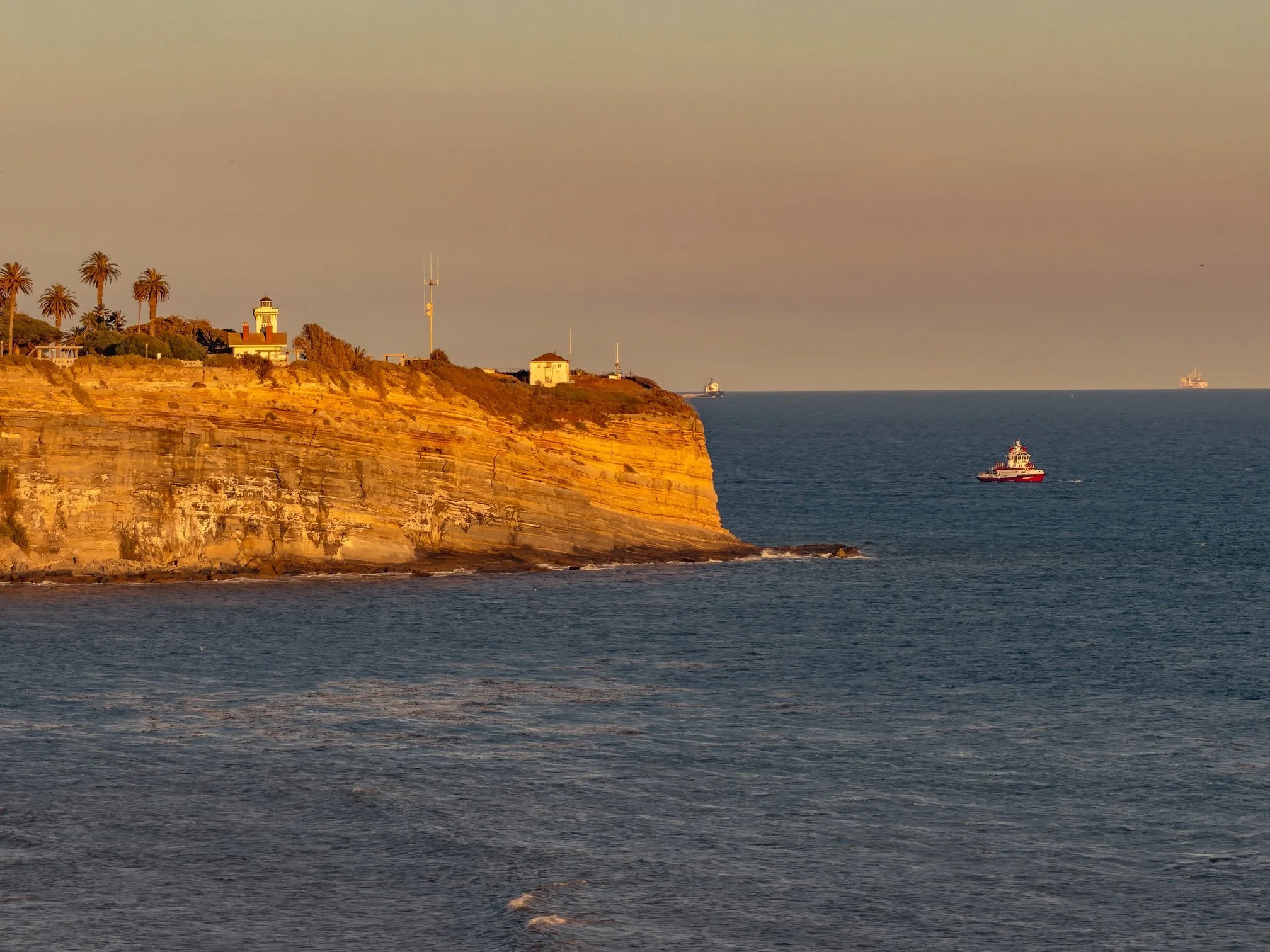 Sunset view of Point Fermin with a cliff, a lighthouse, trees, and several ships on the ocean in the distance.