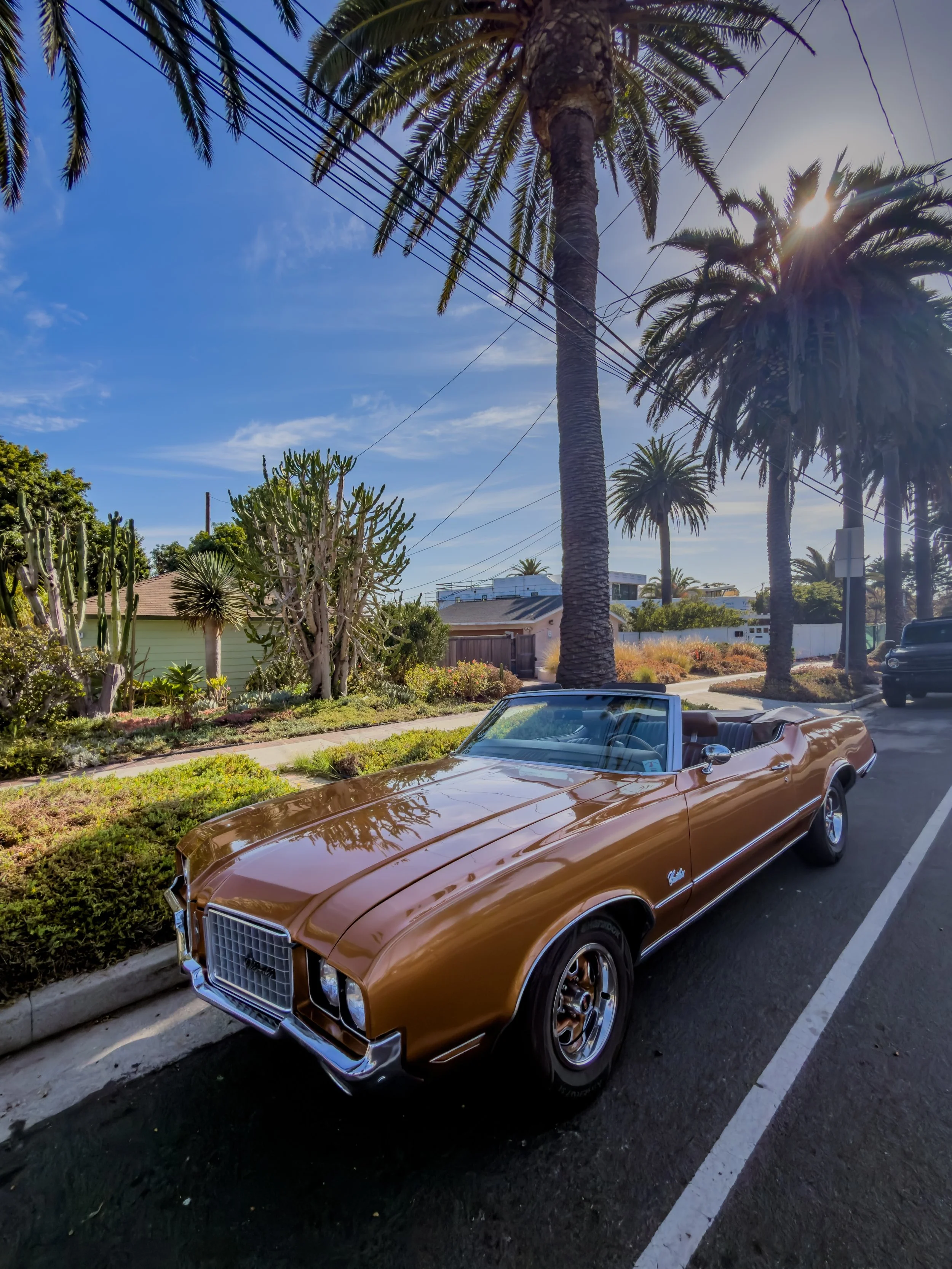 A vintage brown convertible car parked on the side of a street with palm trees and a clear blue sky in the background.