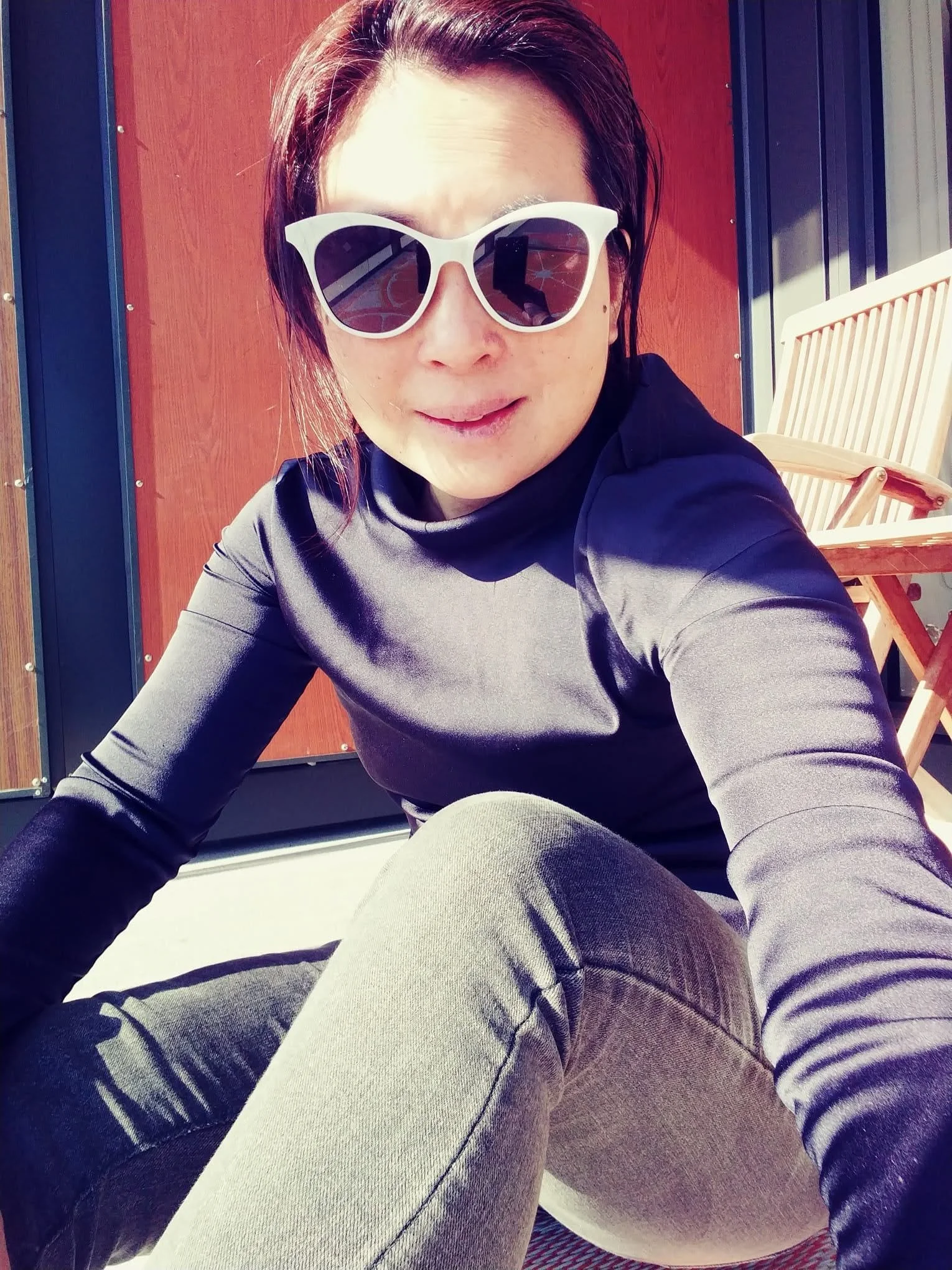 A person with dark hair wearing white sunglasses and a black top taking a selfie outdoors, with a red wooden wall and a chair in the background.