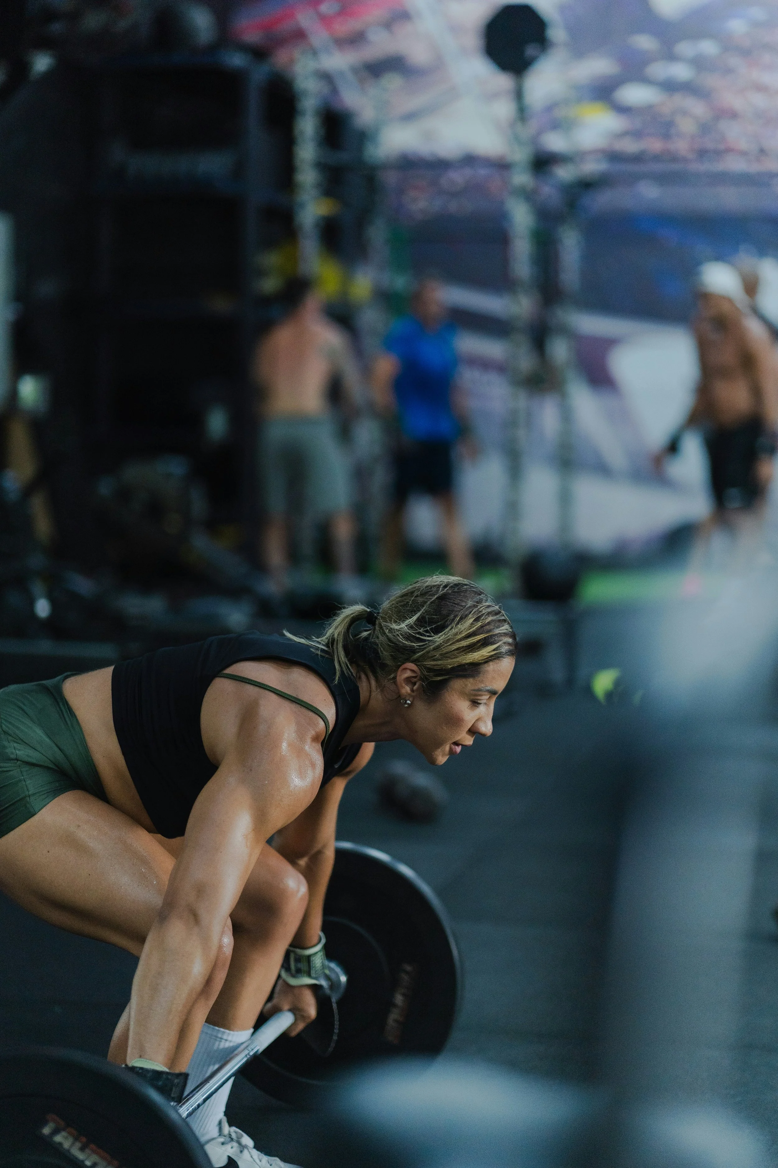 A woman with blonde hair tied back, wearing a black athletic tank top and green shorts, is performing a deadlift in a gym. She is bent over with a barbell loaded with black weights on the floor in front of her. In the background, there is a blurred wall with some people, fitness equipment, and a large mirror.