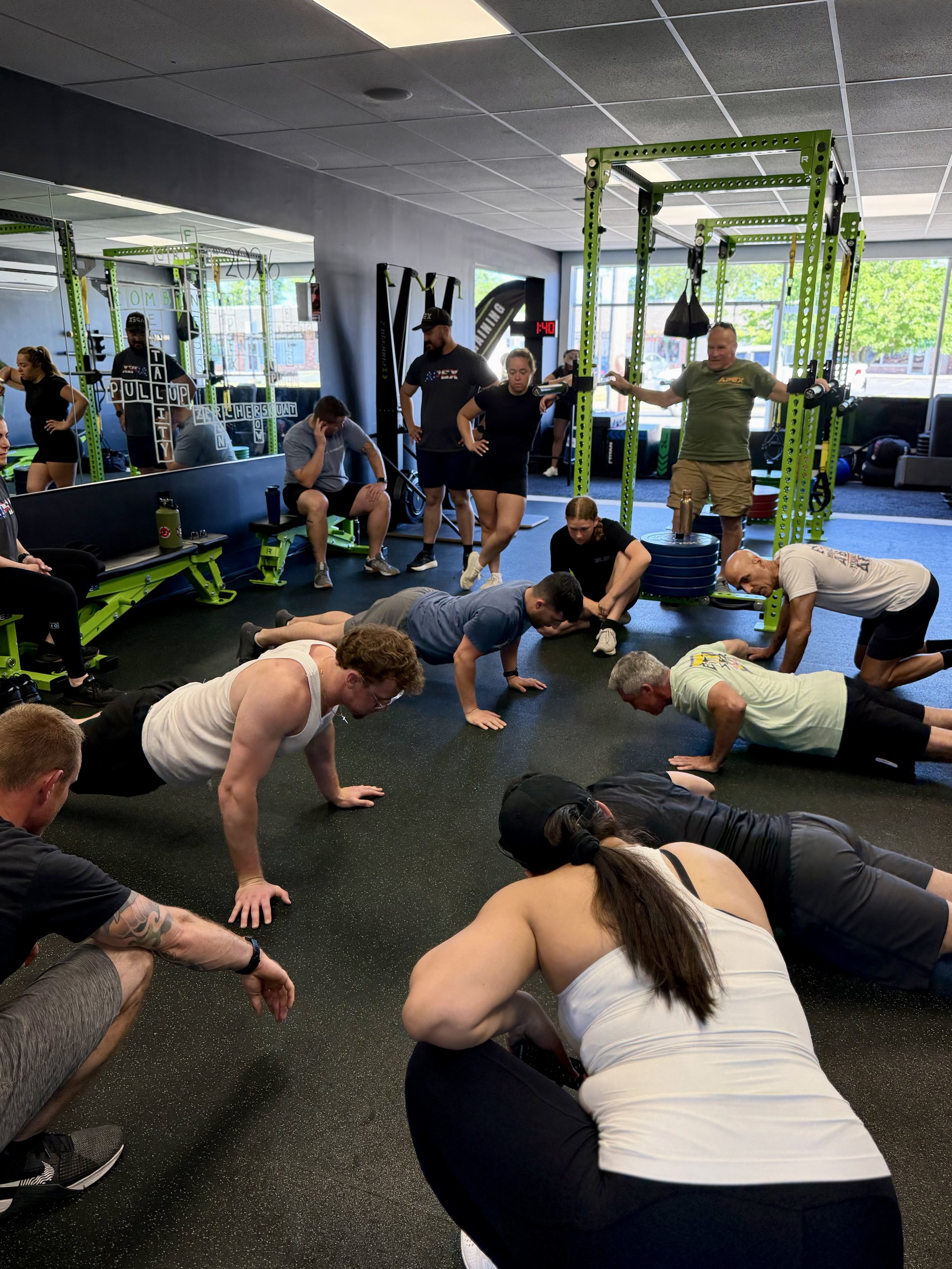 People participating in a fitness class in a gym, performing push-ups on the floor with some sitting and standing nearby.