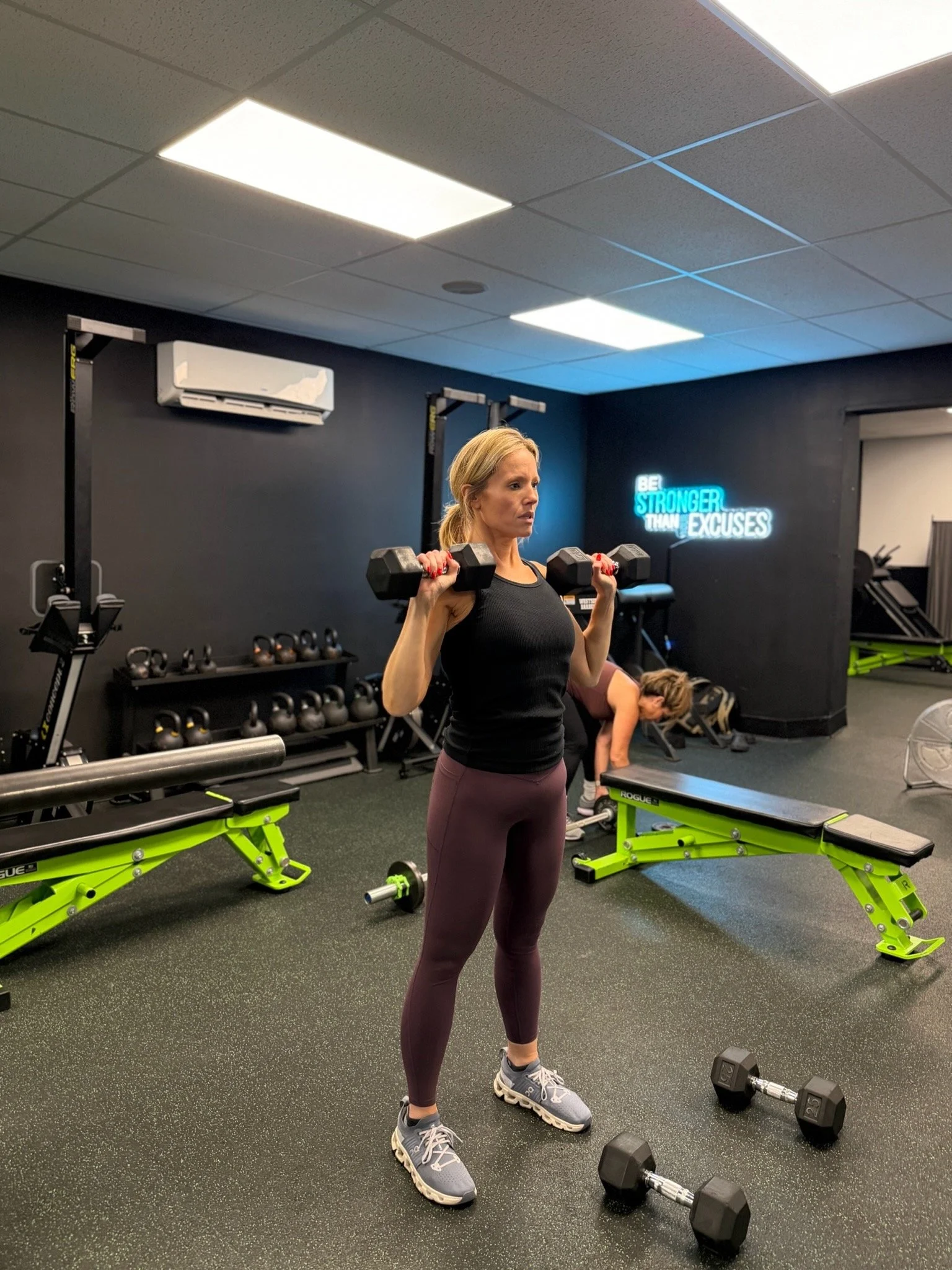 A woman with blonde hair, wearing a black tank top and maroon leggings, lifting dumbbells in a gym. There are fitness equipment and a motivational sign on the wall that reads "Be Stronger Than Your Excuses."