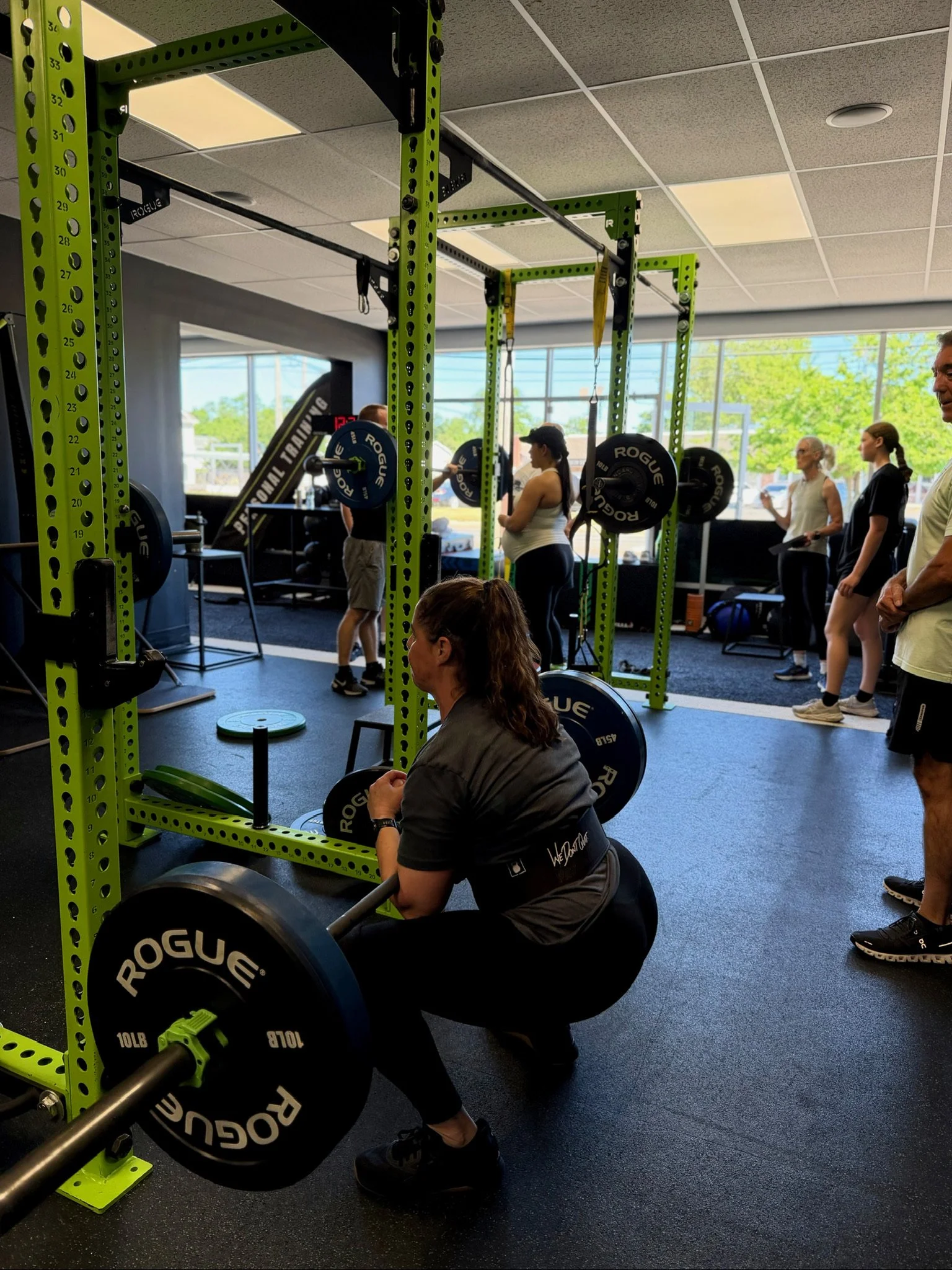 A woman in black workout clothes lifting a barbell with weight plates in a gym. Other people are in the background, some standing and talking. The gym has large windows and a ceiling with grid-style tiles.