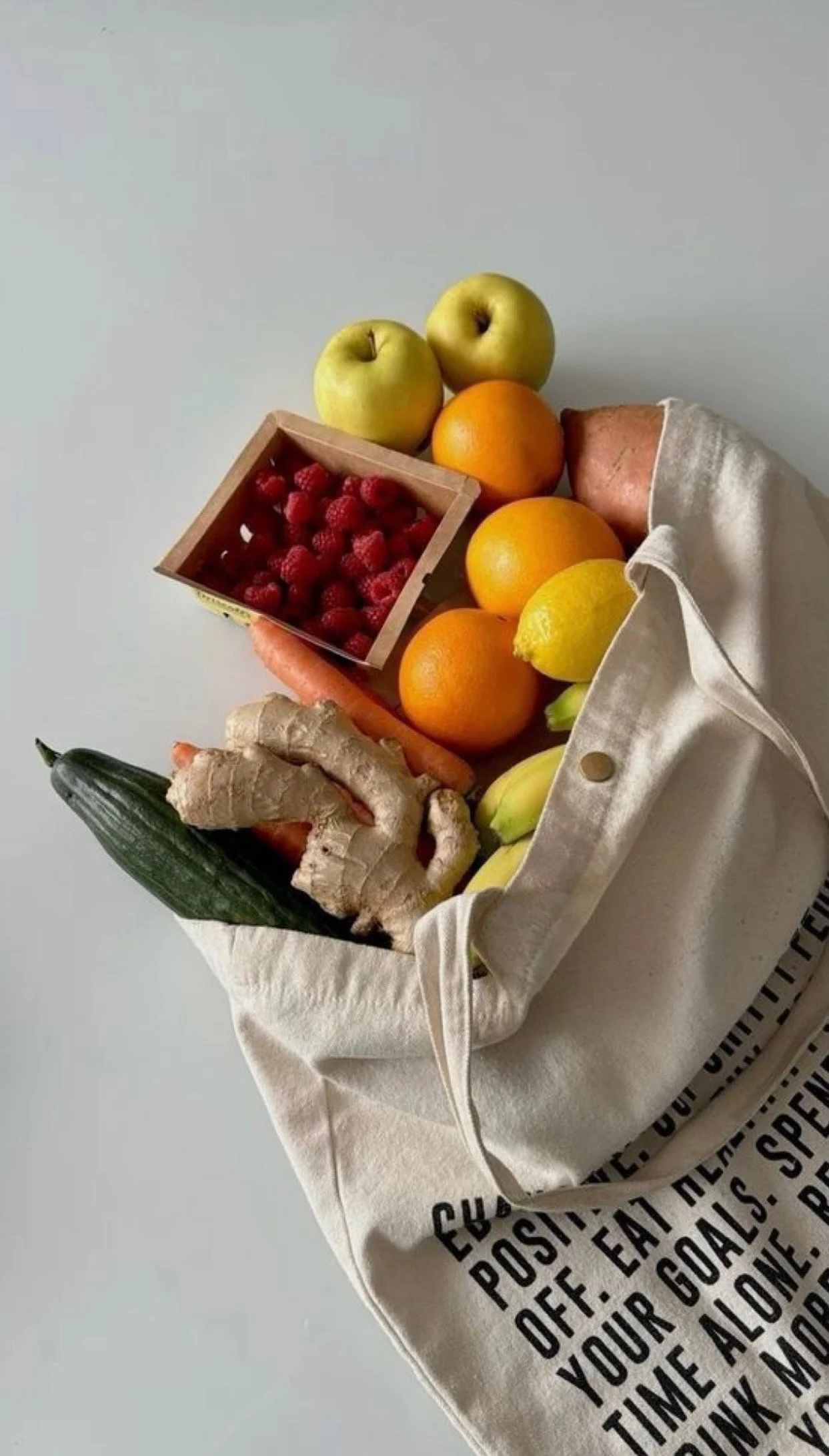 A collection of fresh fruits including apples, oranges, lemon, cucumber, carrot, ginger, and a box of raspberries arranged on a white cloth and cardboard box.