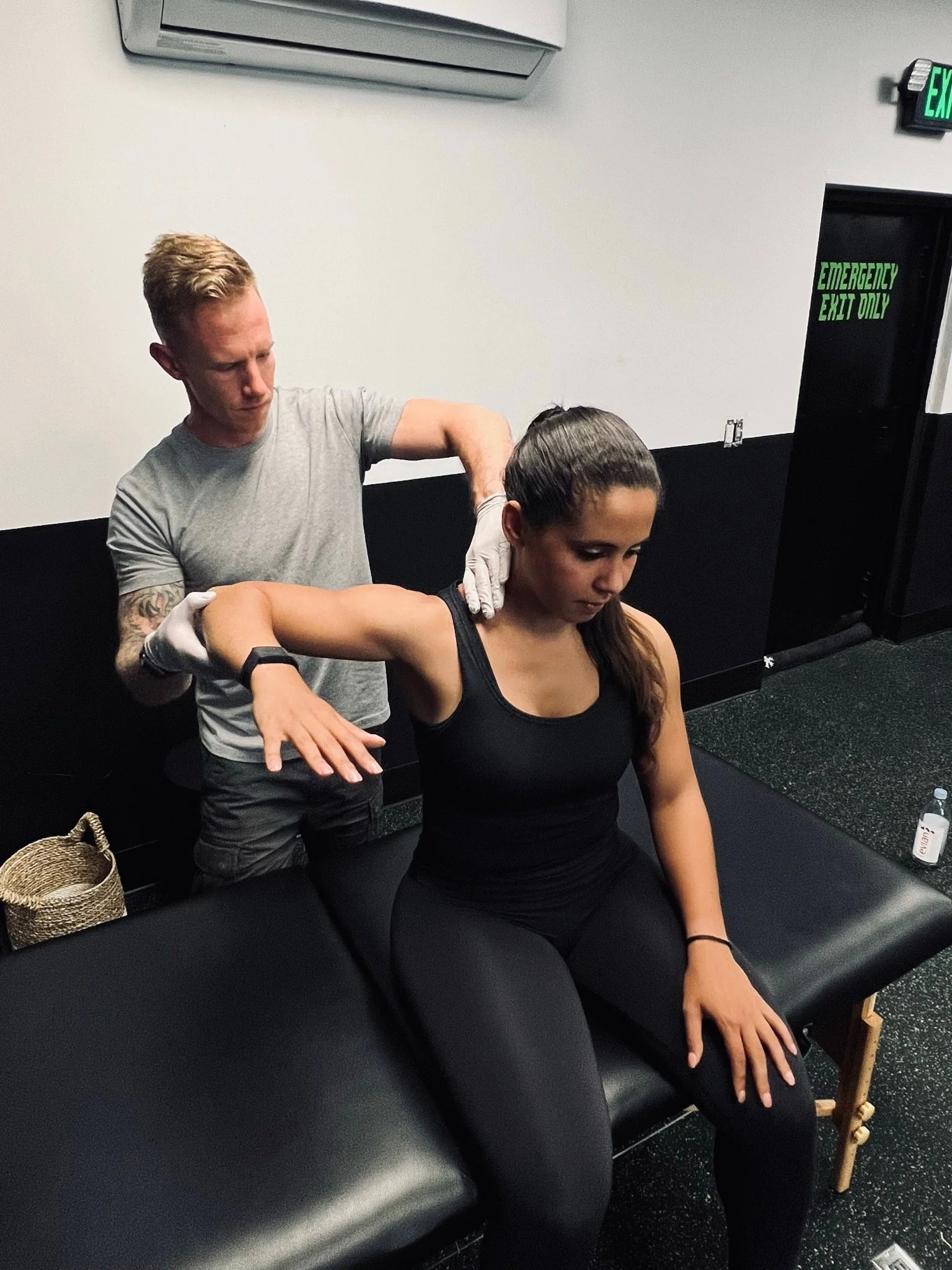 A man with tattoos on his arm and wearing a gray t-shirt is massaging a woman with long dark hair who is seated with her left arm extended and wearing a black tank top. They are in a room with white and black walls, an air conditioning unit, an emergency exit sign, a water bottle, a woven bag, and a wooden chair.