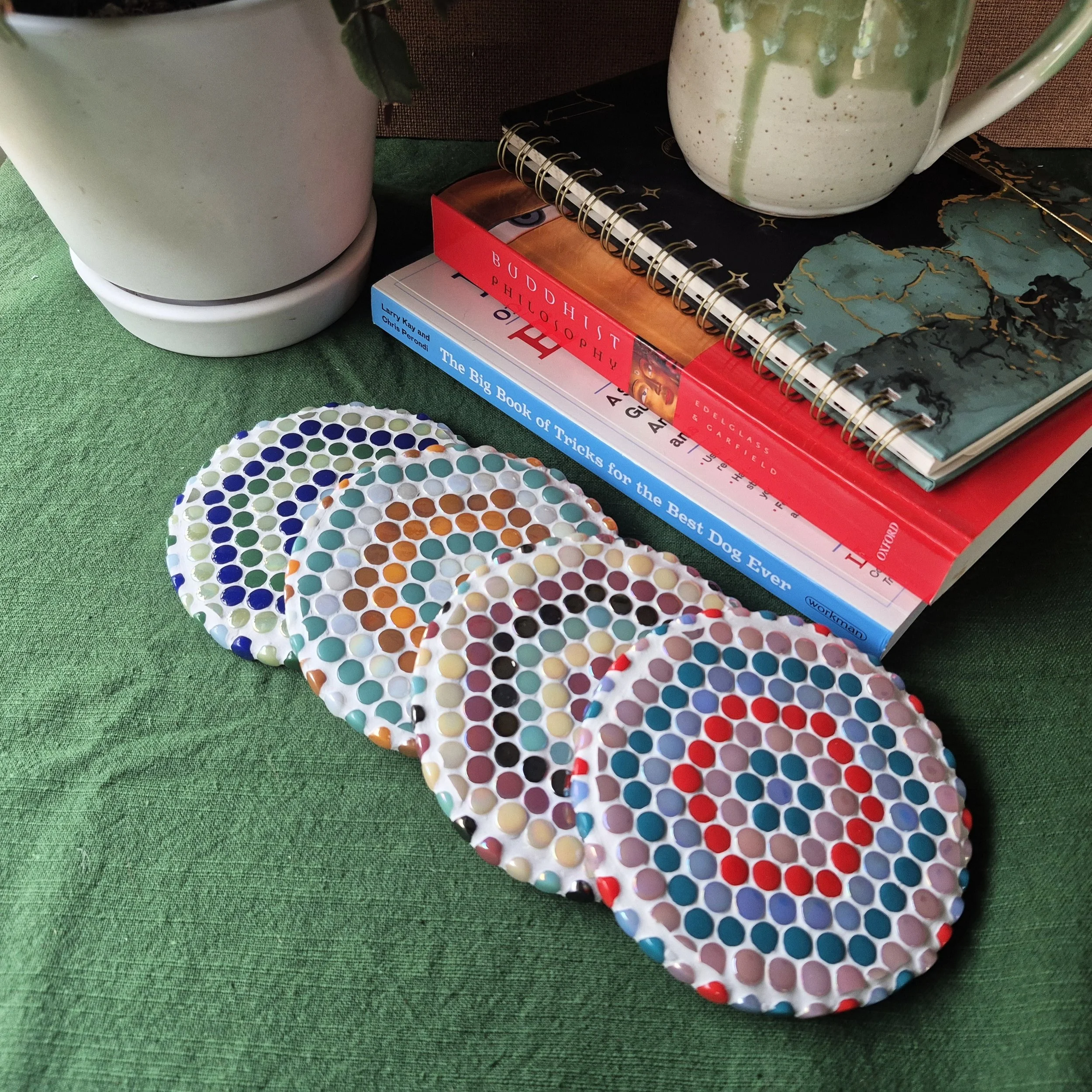Close-up of decorative ceramic plates with colorful dots, stacked on green textured fabric, alongside books and mugs with pottery designs, and a white ceramic container.