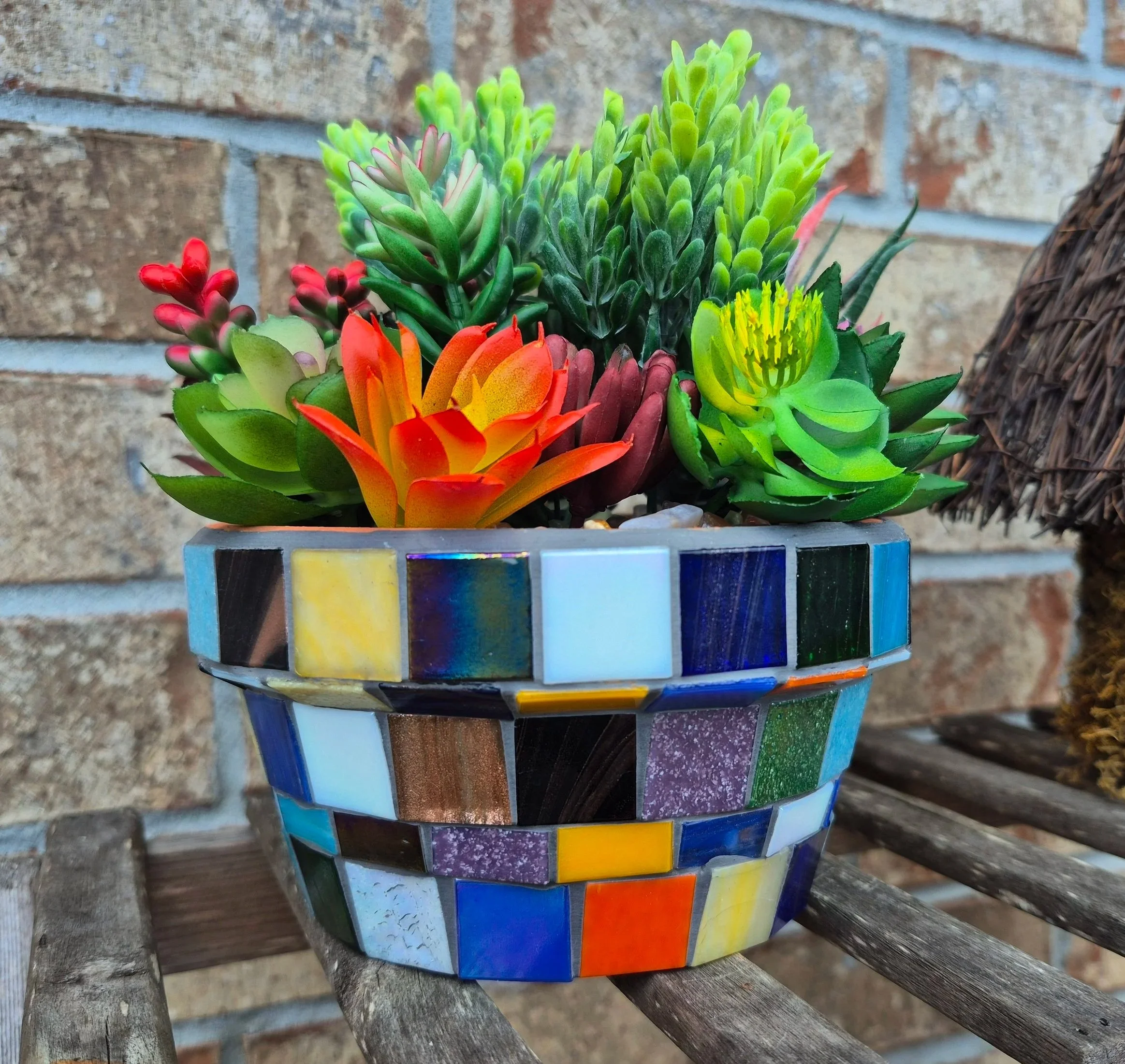 Colorful mosaic planter holding assorted succulents in front of a brick wall.