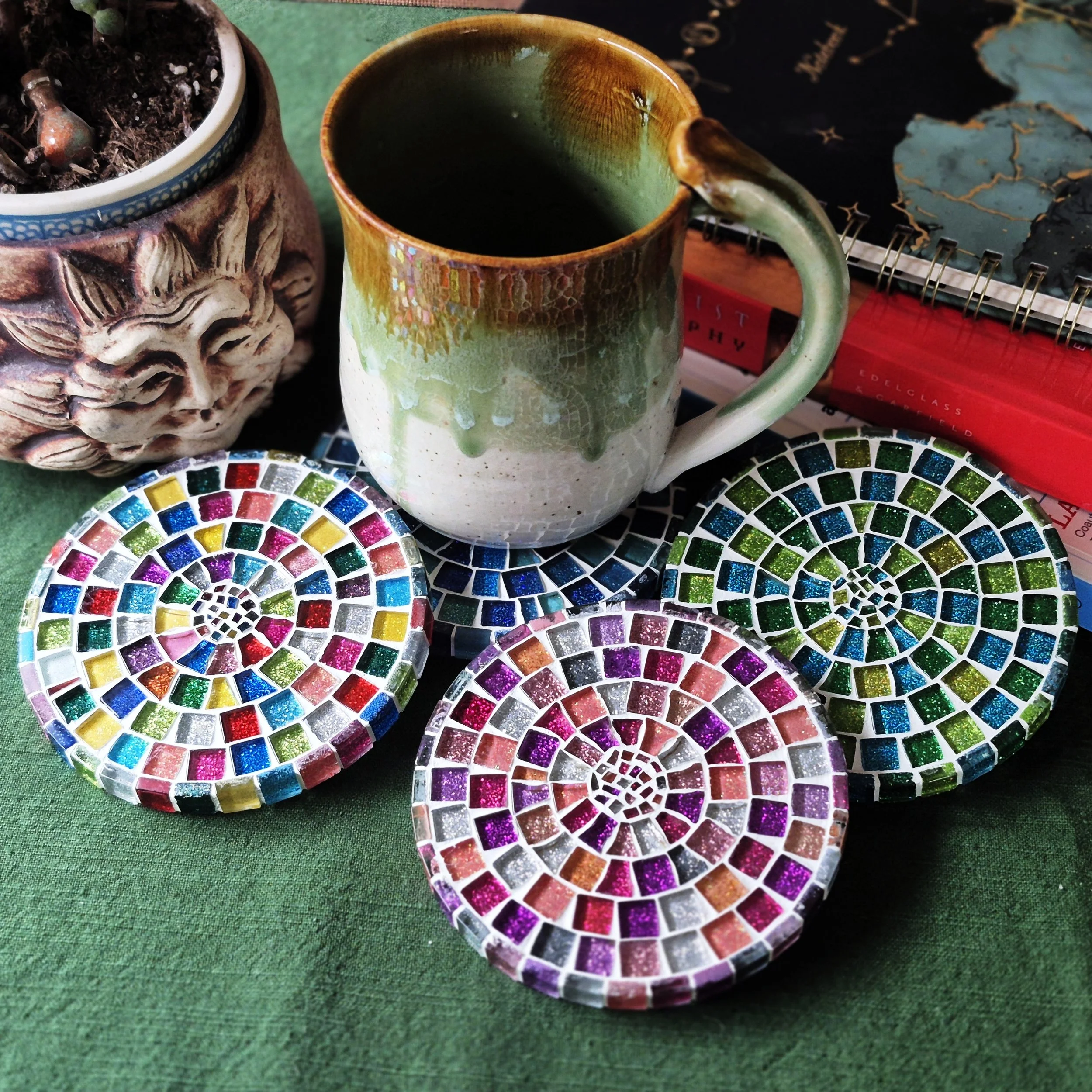 Colorful mosaic coasters featuring spiraling patterns in vibrant colors such as purple, pink, blue, green, and yellow, placed on a green surface. A ceramic mug with a gradient glaze from white to green and brown, containing a dark liquid, is also visible along with a potted plant and some books in the background.