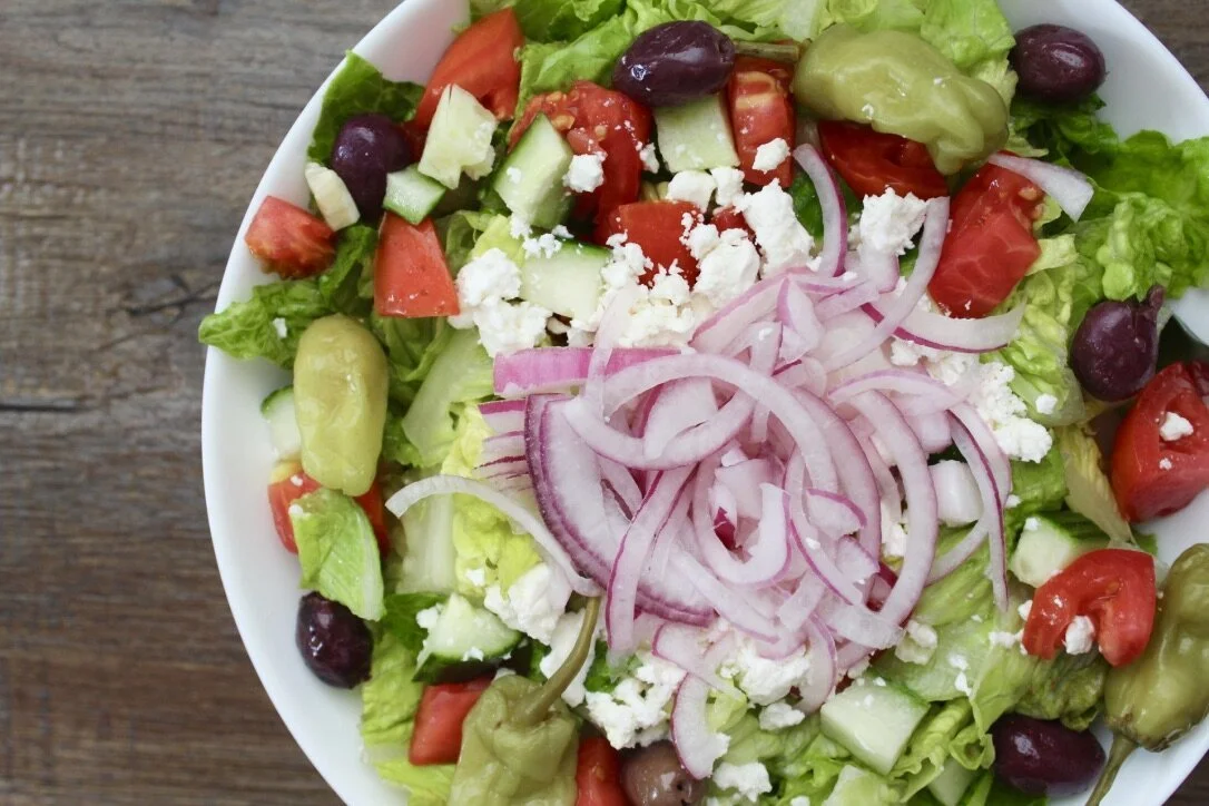 Mixed green salad with tomatoes, cucumbers, red onions, black olives, green olives, and crumbled feta cheese in a white bowl on a wooden surface.