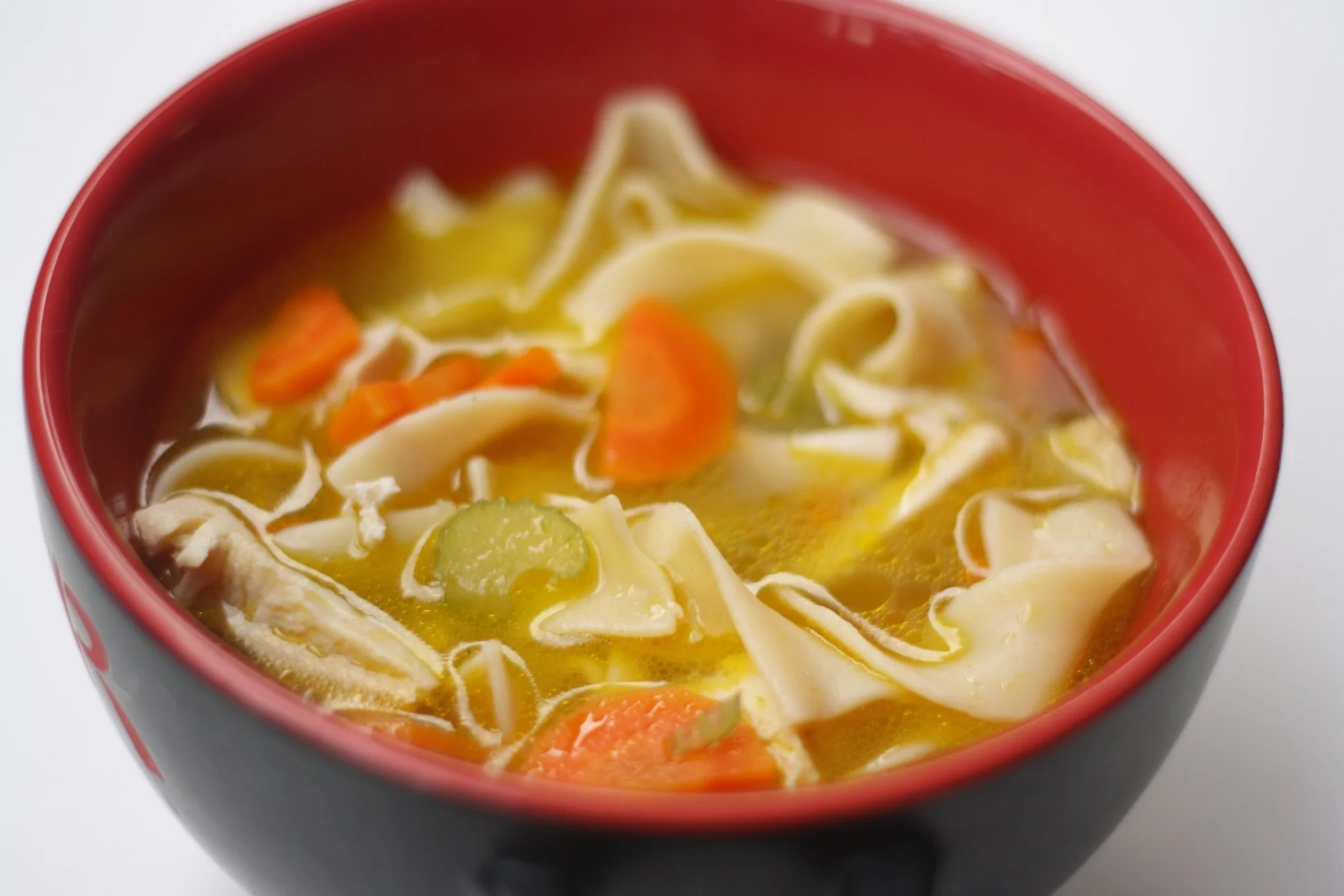 A cup of vegetable noodle soup containing sliced carrots, zucchini, shredded chicken, and noodles in a yellow broth, served in a red and black bowl.
