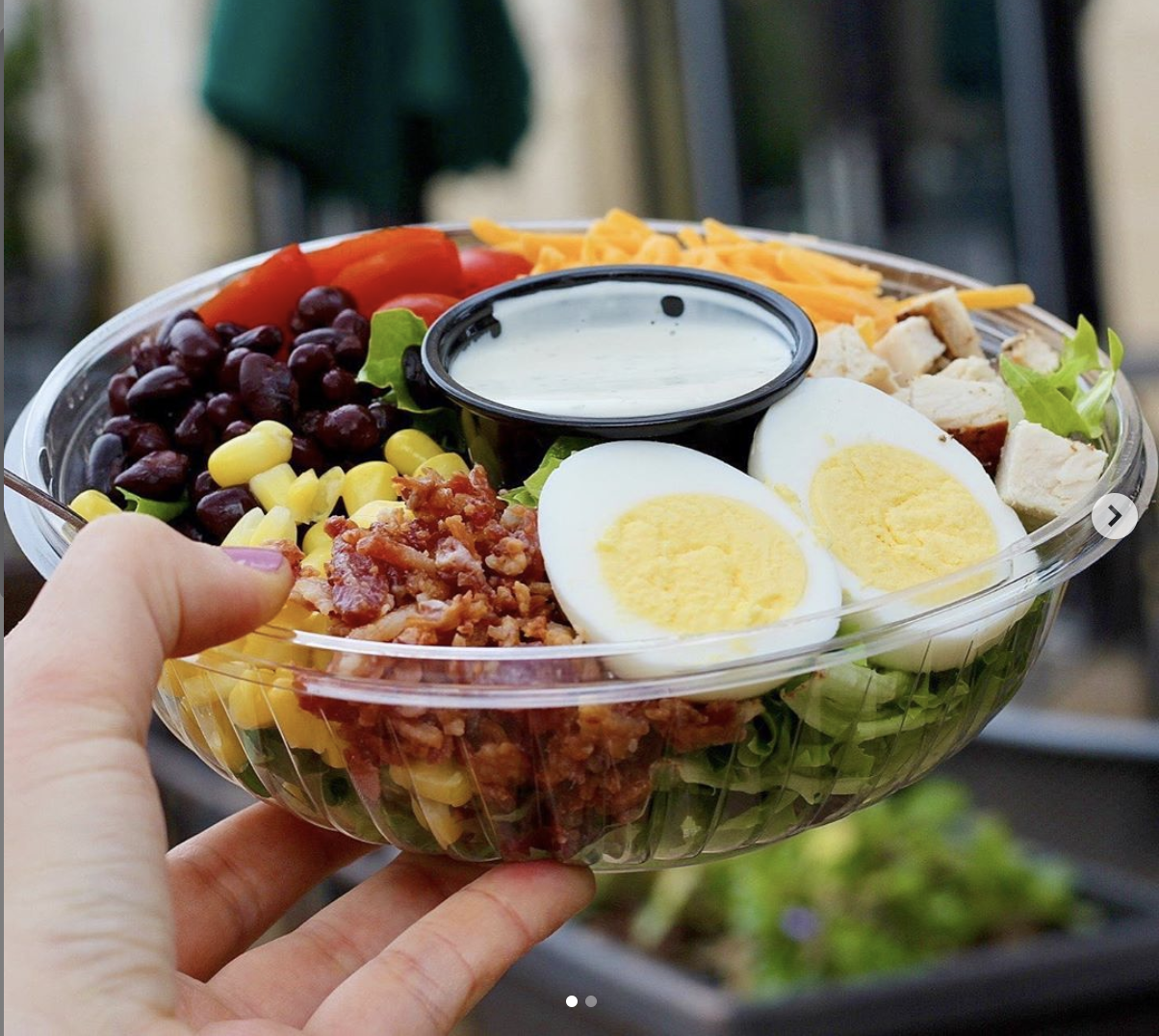A hand holding a large plastic bowl of salad with various ingredients including grapes, corn, chopped chicken, half a boiled egg, shredded cheese, cherry tomatoes, lettuce, and a small container of dressing in the center.
