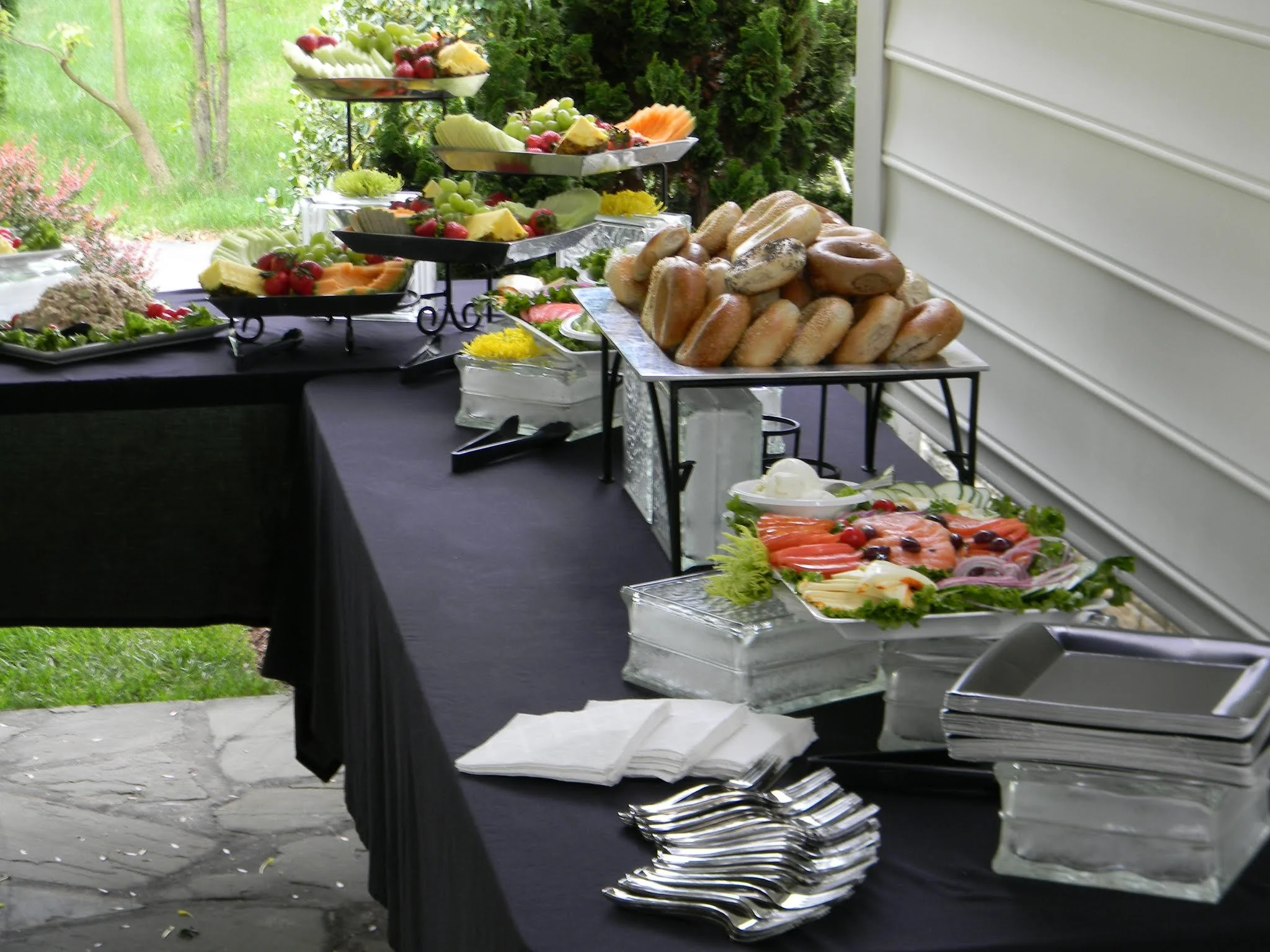 Buffet table outdoors with trays and stands filled with fresh fruit, vegetables, bagels, smoked salmon, and salad, set up in a residential backyard.