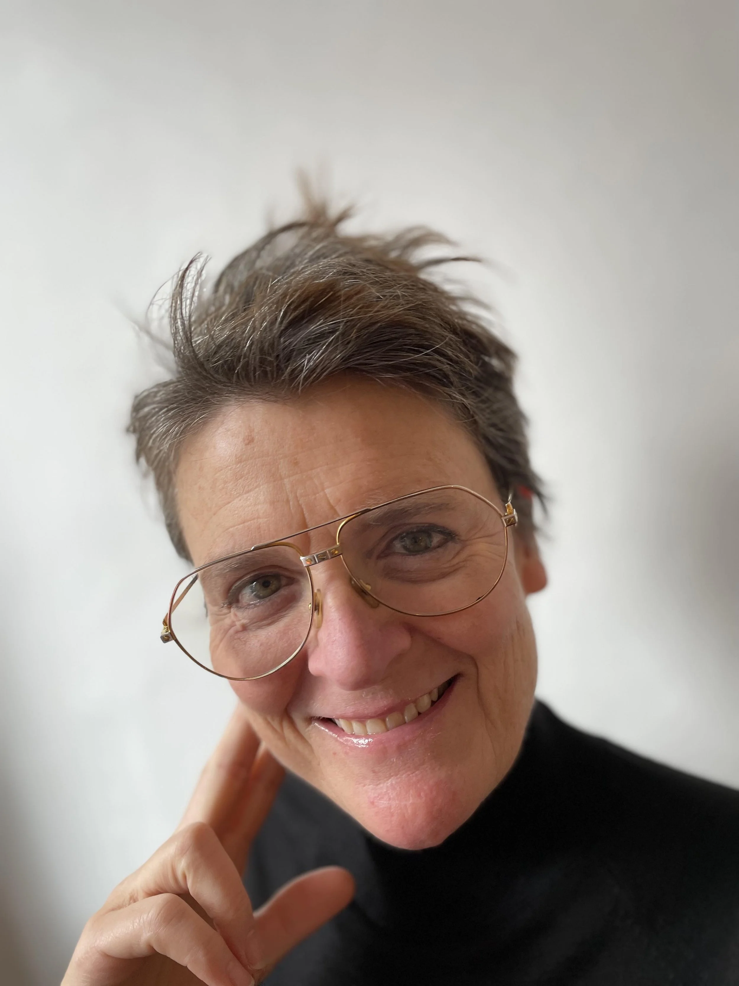 A woman smiling, wearing glasses, with short brown hair, touching her face with her left hand.
