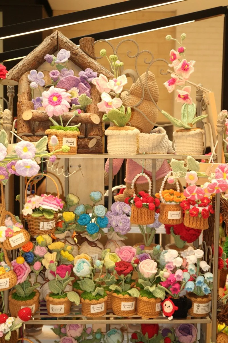 Display of colorful crocheted flowers and plants in baskets and pots, arranged on a metal stand and wooden shelf.