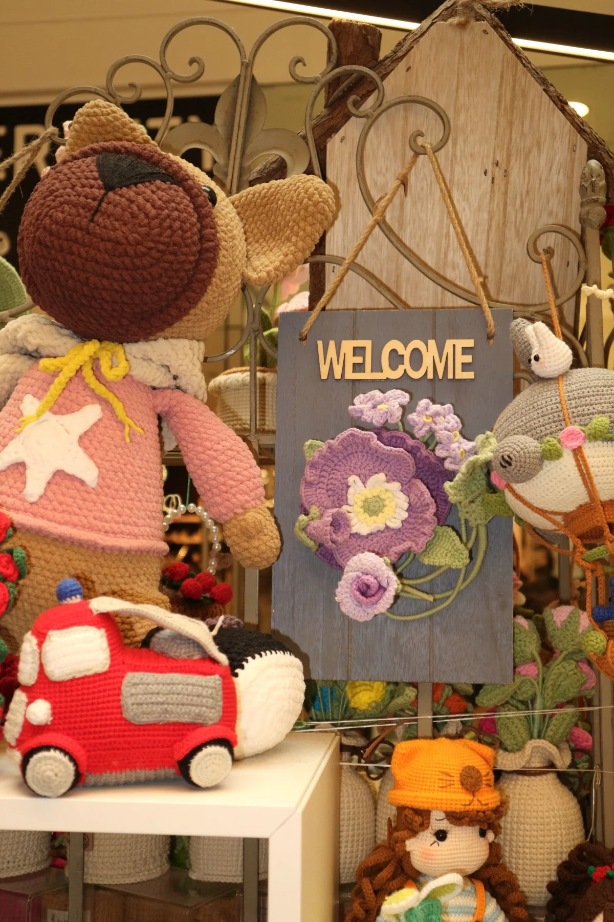 Display of various handmade crochet stuffed animals and decorative items, including a large animal doll wearing pink with a star on its chest, a red and white toy fire truck, and a welcome sign with embroidered purple flowers.