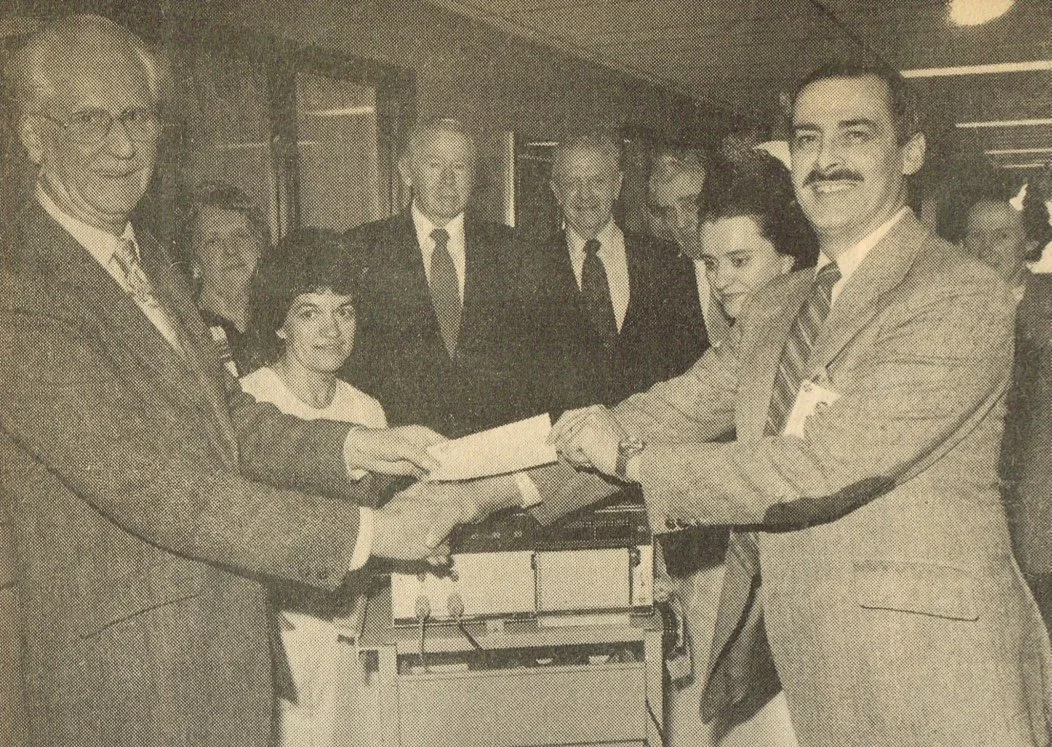 A group of people, including men in suits and a woman, gathered around a table where two men are shaking hands and exchanging a document, in a formal setting.