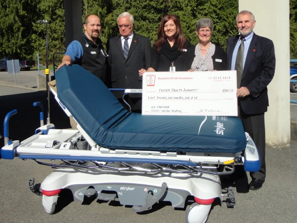 Five people stand outdoors behind a large medical stretcher with a blue cover. They are holding a giant check, with trees and parked cars in the background. The check is from the Keystone of Life Foundation, made out to Fraser Health Authority, dated July 23, 2015, for $8,905.05.