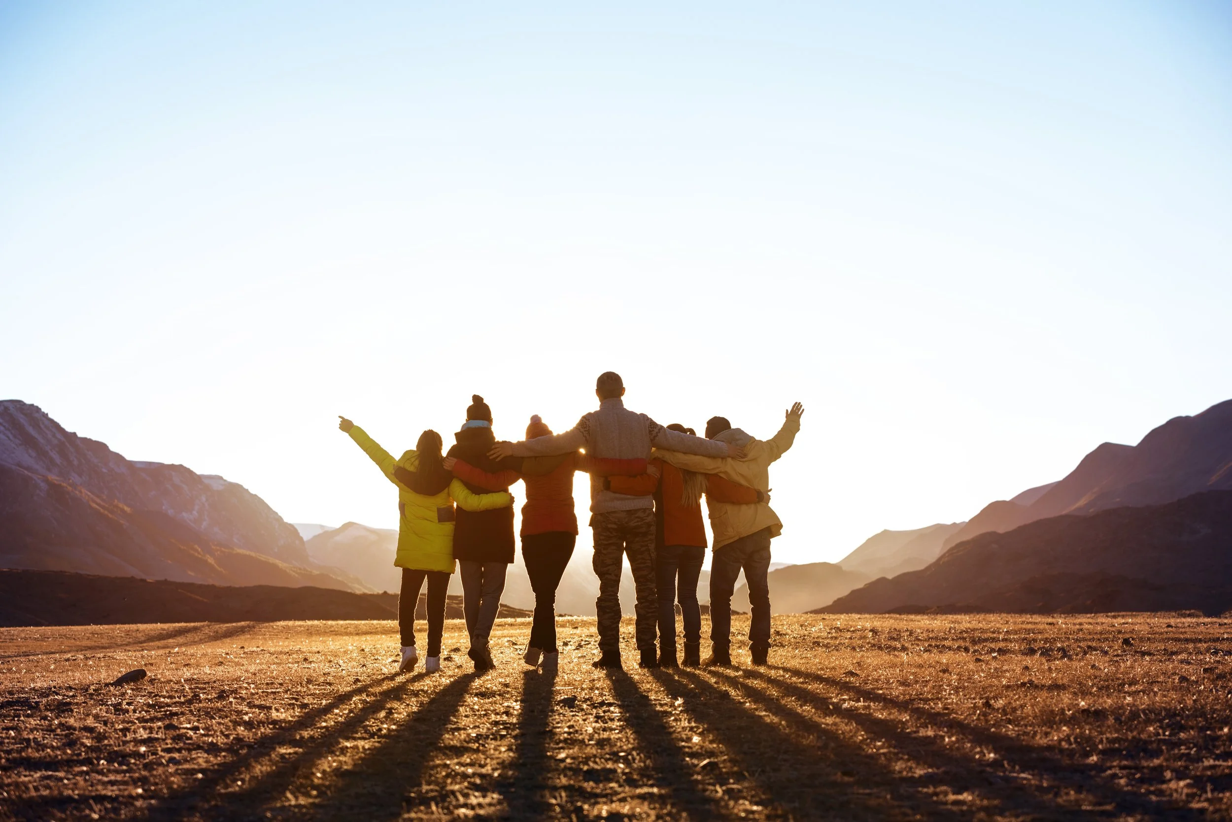 A group of people standing together outdoors in a mountainous area during sunset, with their arms around each other and facing the sunset.